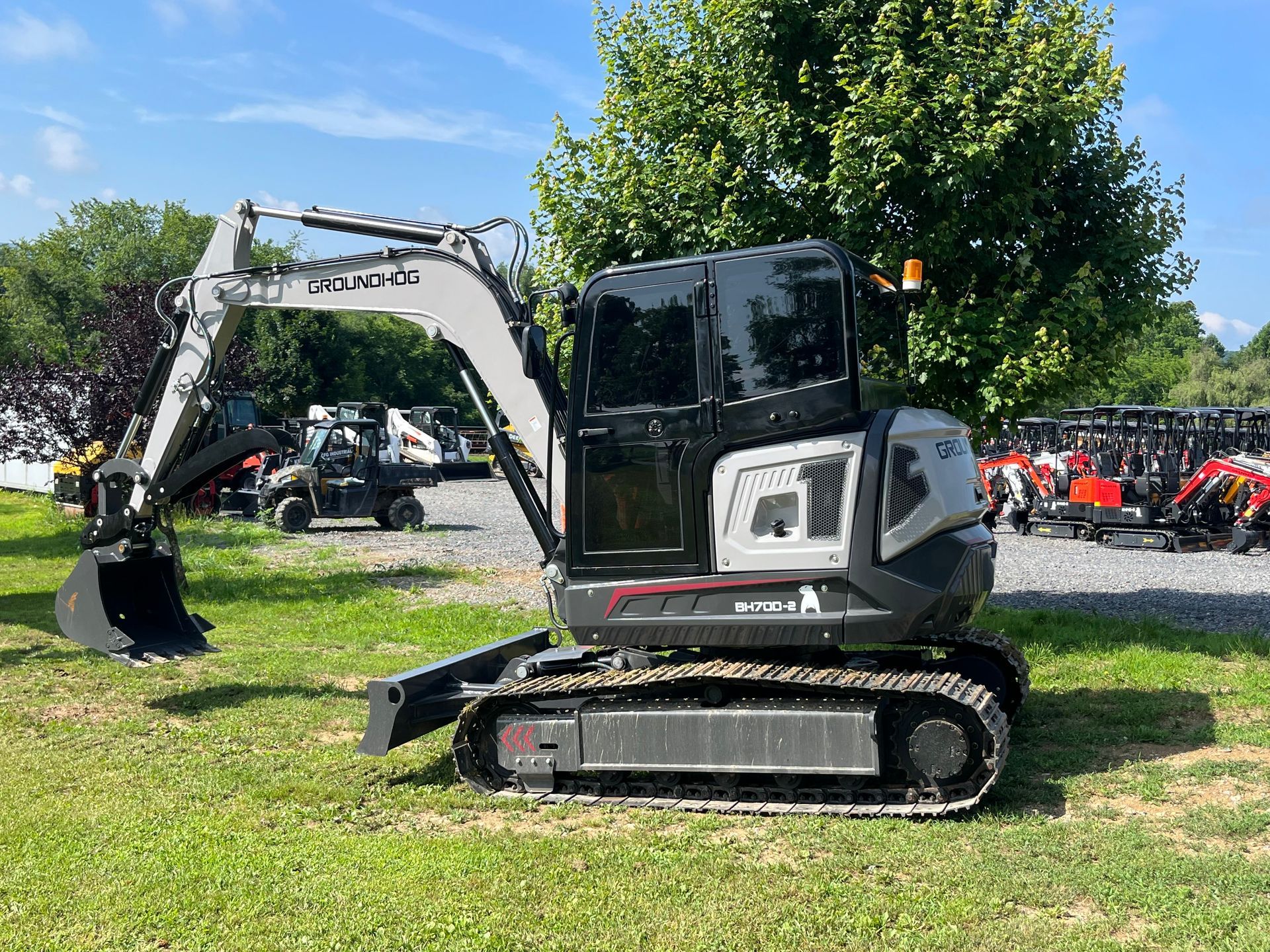 Gray compact excavator on grass at an outdoor equipment display, with red machinery in the background.