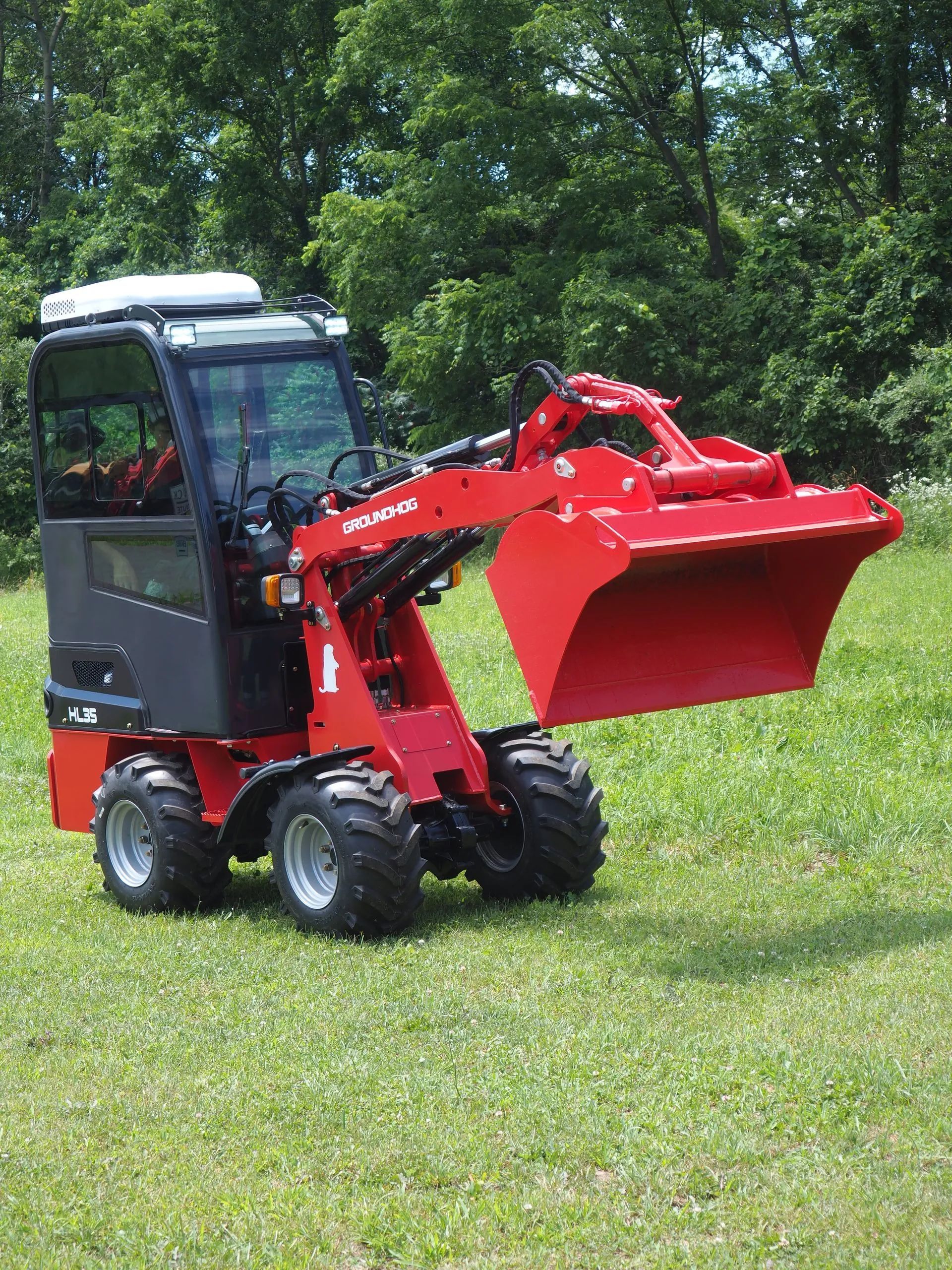 A red compact articulated loader with an enclosed cab sits on a grassy field with its bucket raised.
