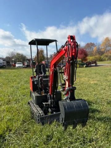 A compact red mini-excavator with a black canopy parked in a grassy field under a bright blue sky.