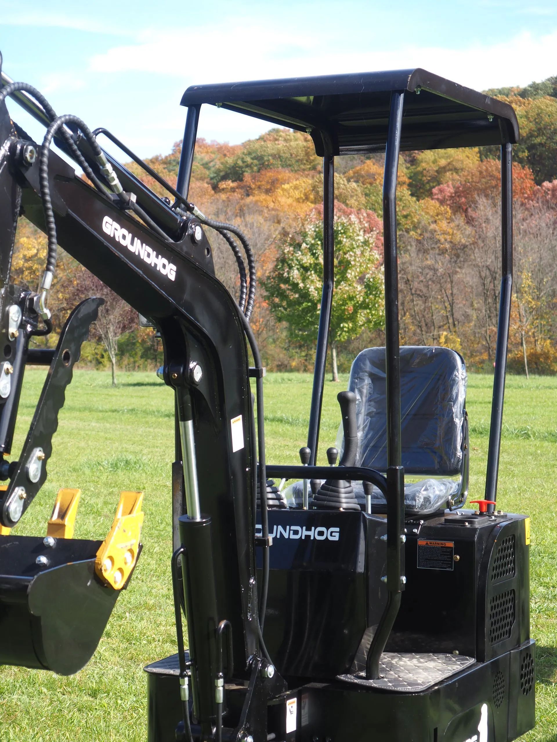 A black Groundhog brand mini excavator with a canopy and seat, parked on a grassy field with autumn trees in the background.