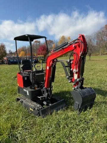 A red and black compact excavator sits on a grassy field under a sunny, partly cloudy sky.