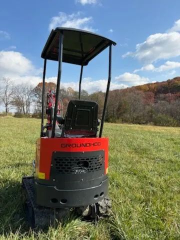 A red and black GroundHog mini-excavator with a canopy, parked in a grassy field under a blue sky.