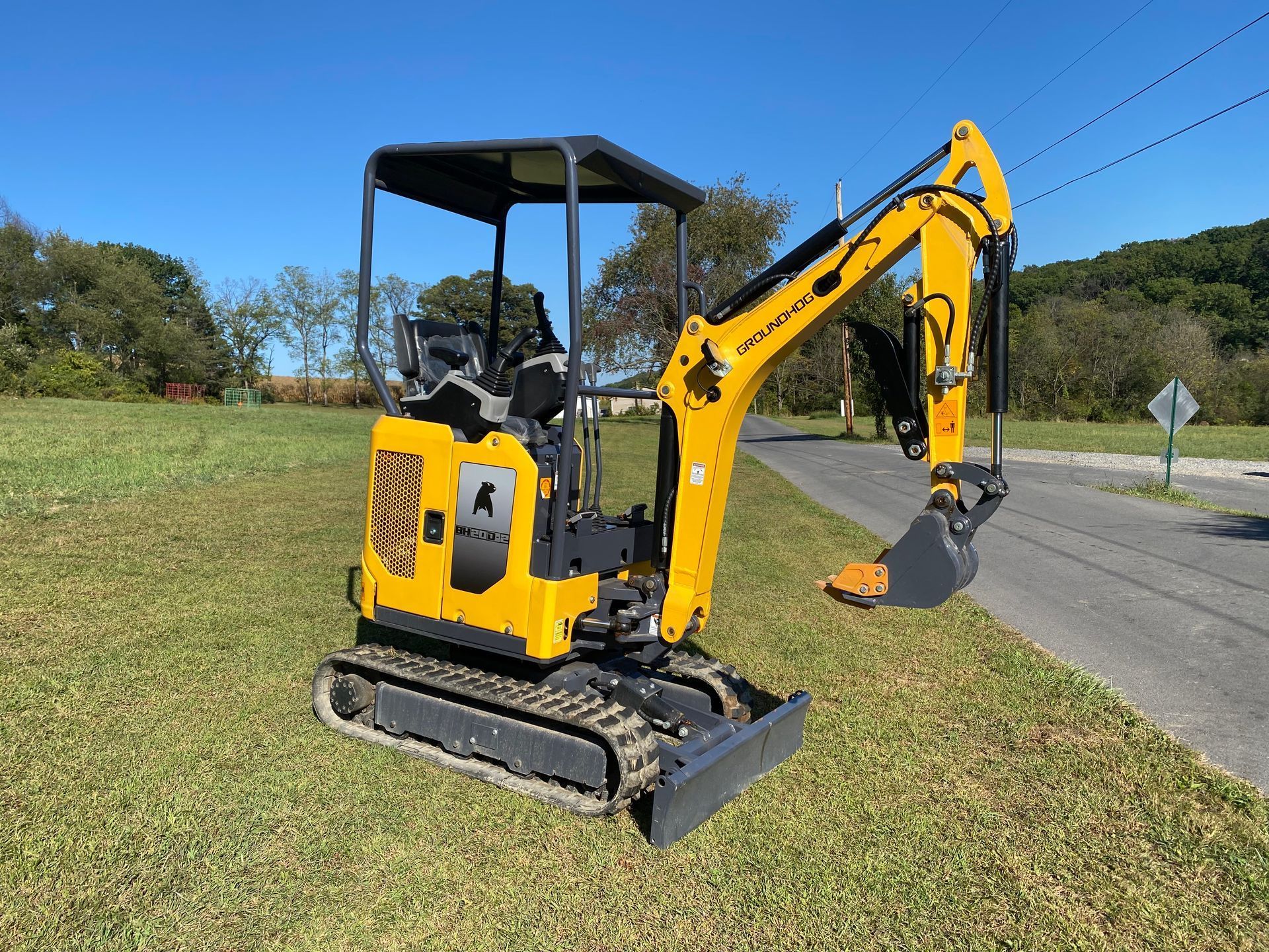 A yellow compact excavator parked on a grassy roadside under a clear blue sky.