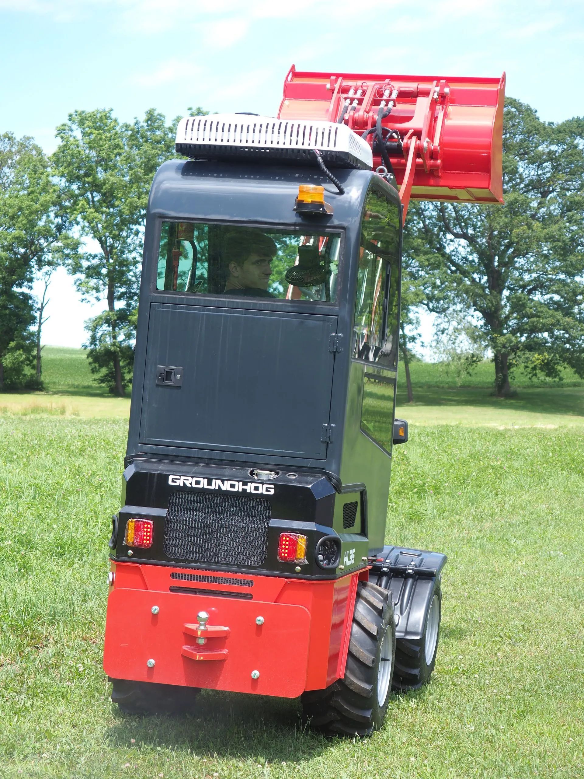 A red and black Groundhog compact utility tractor with a raised front loader bucket parked in a grassy field.