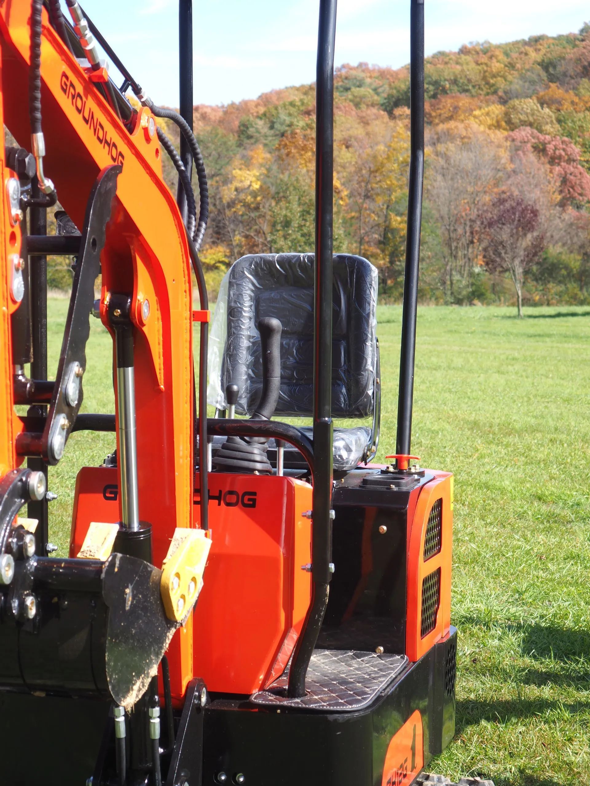 An orange mini-excavator sits in a grassy field with autumn trees in the background.