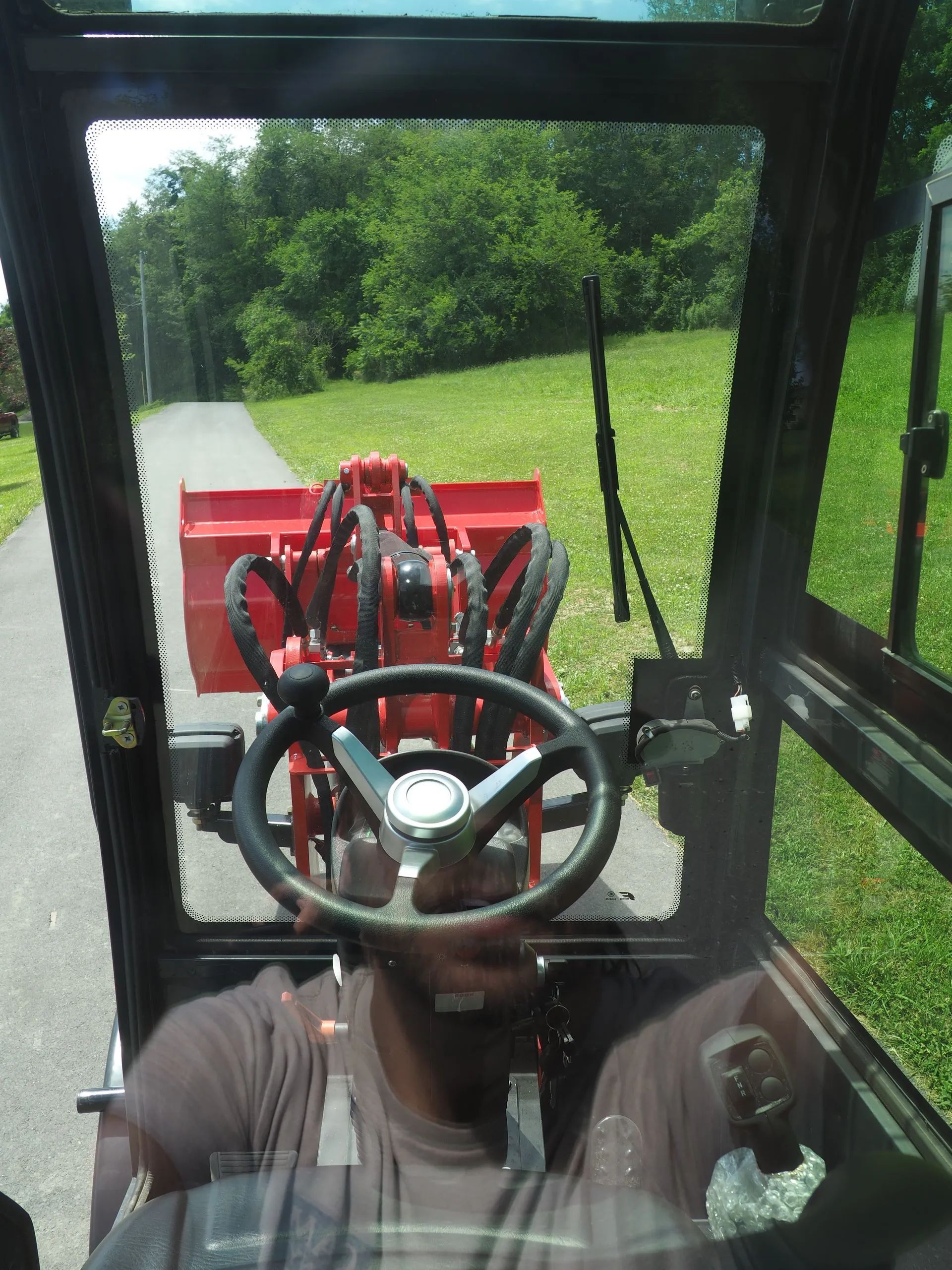 A driver’s view from inside a tractor cabin, looking through the steering wheel at a red loader bucket on a paved road.