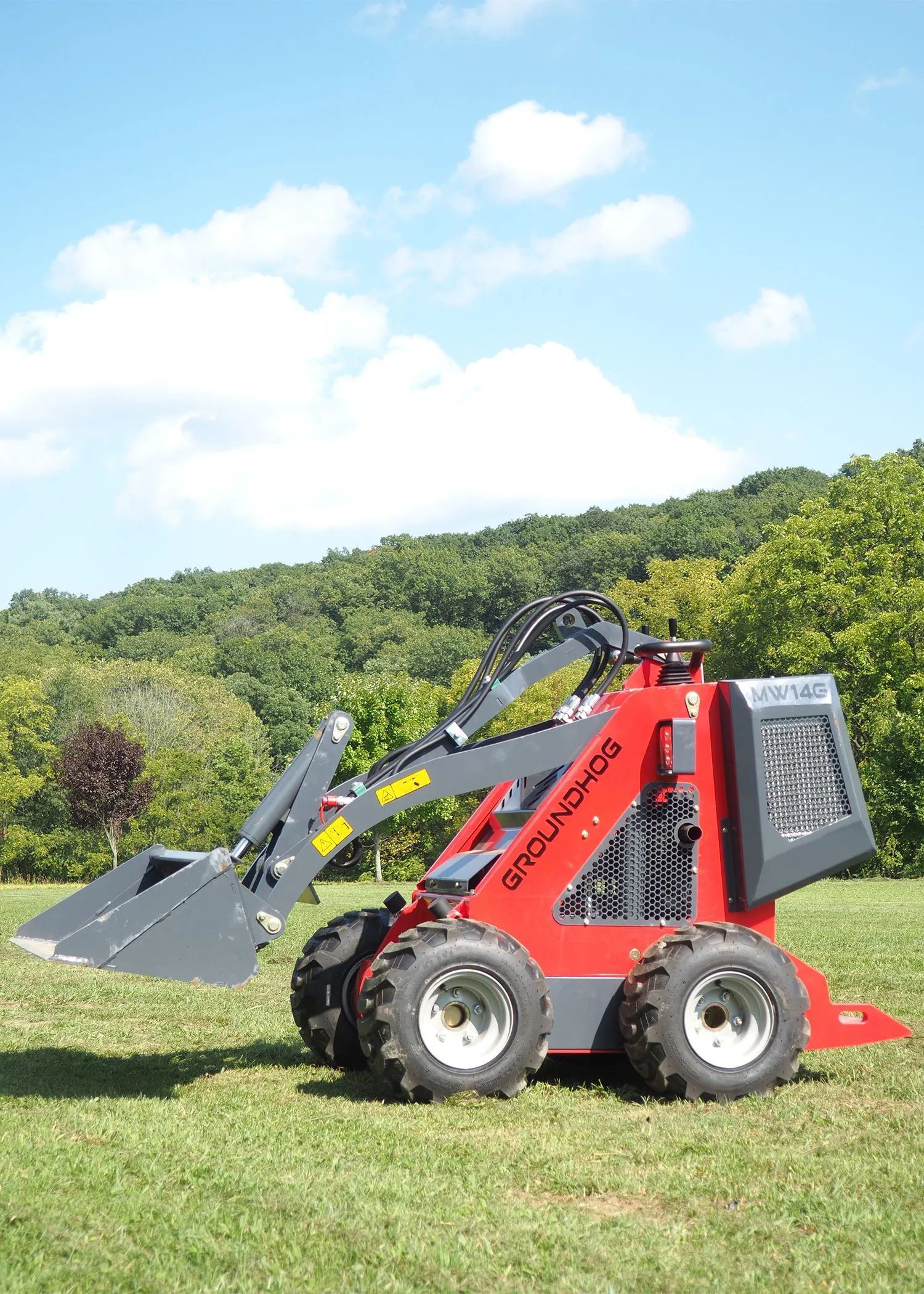 A red skid steer loader with a grey bucket sits on a grassy field against a backdrop of trees and a blue sky.