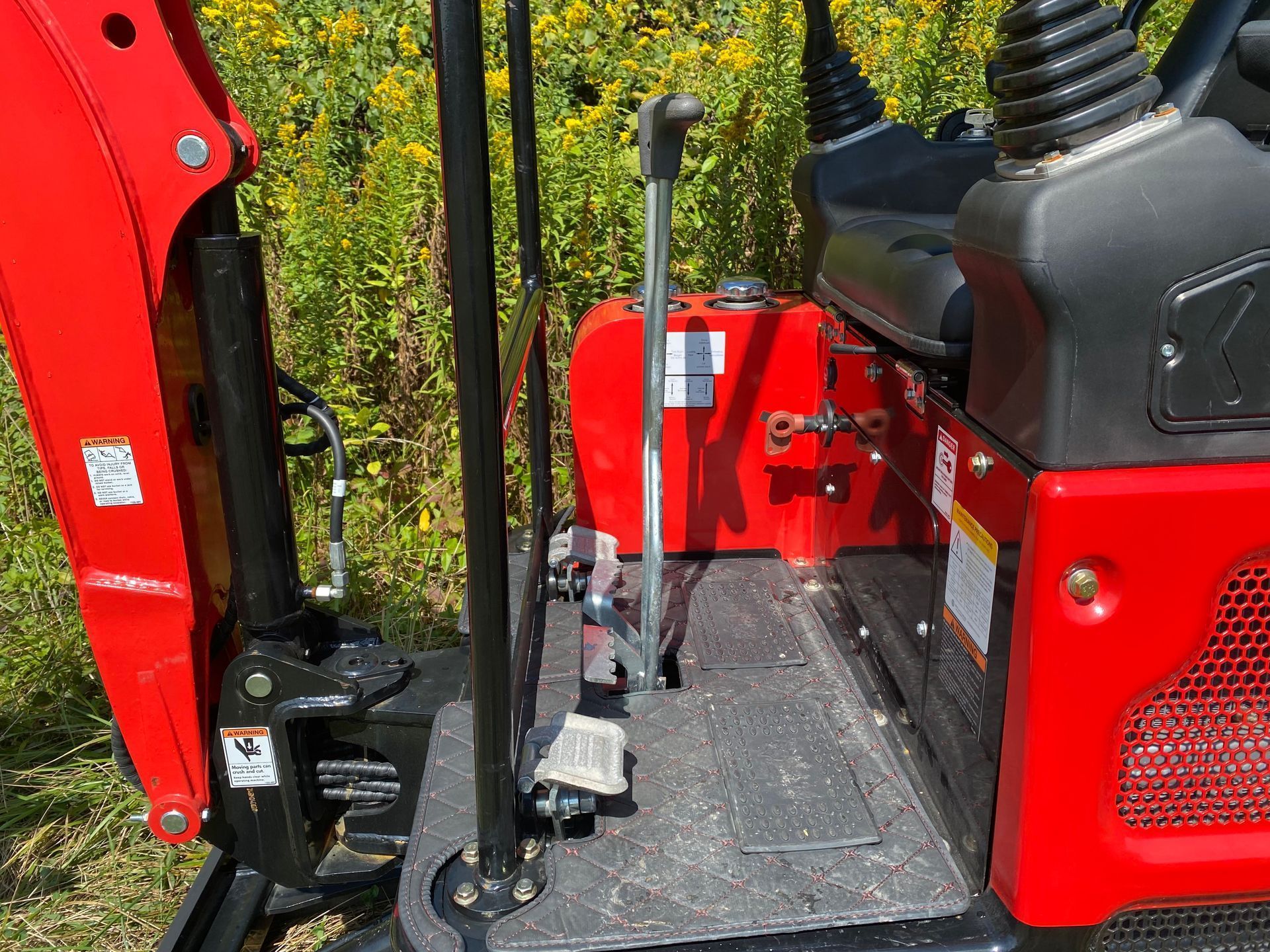 Close-up of the operator platform of a red mini-excavator, showing a control lever and pedals amidst outdoor foliage.
