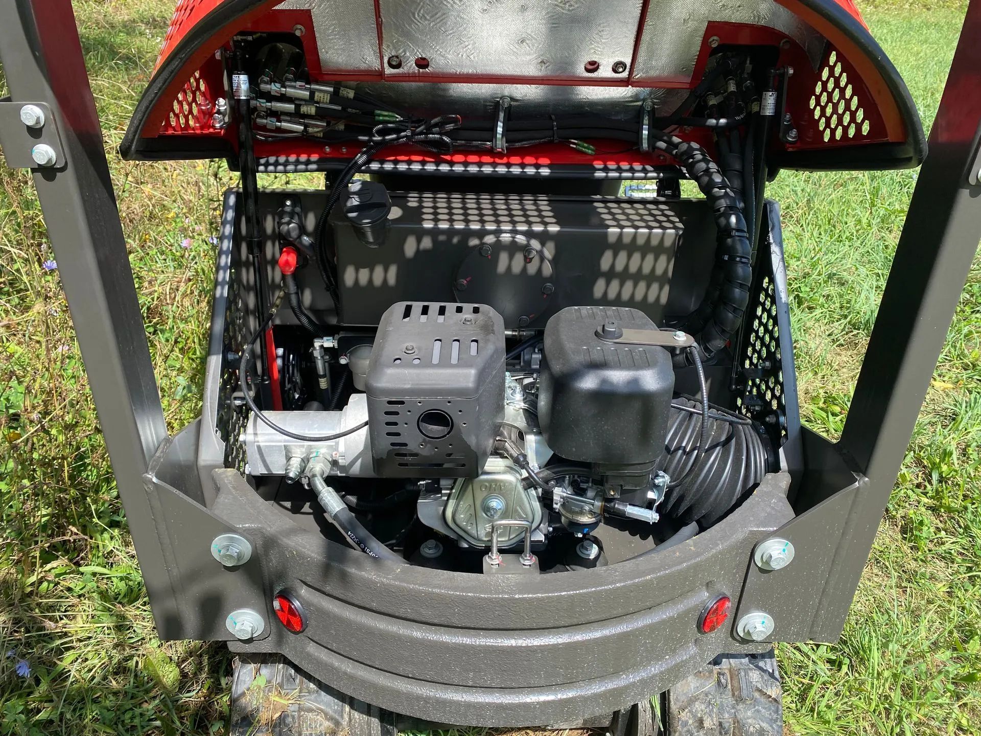 The rear view of a small red construction machine showing its engine, battery, and cooling fans in a grassy field.