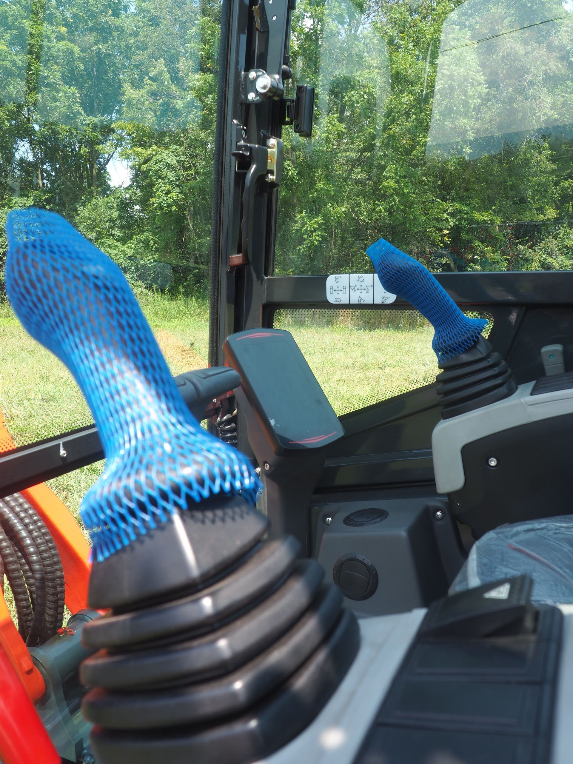 Two black joysticks inside an equipment cabin, both covered in protective blue mesh netting, with trees visible outside.