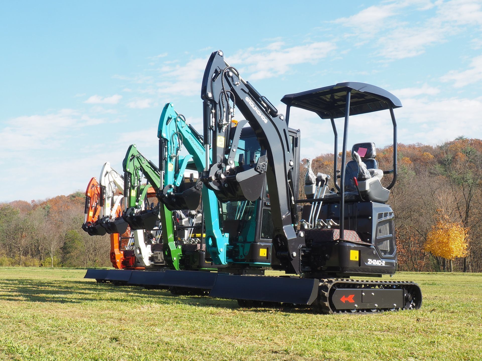 Five mini excavators in orange, white, green, teal, and black lined up in a grassy field under a blue sky.