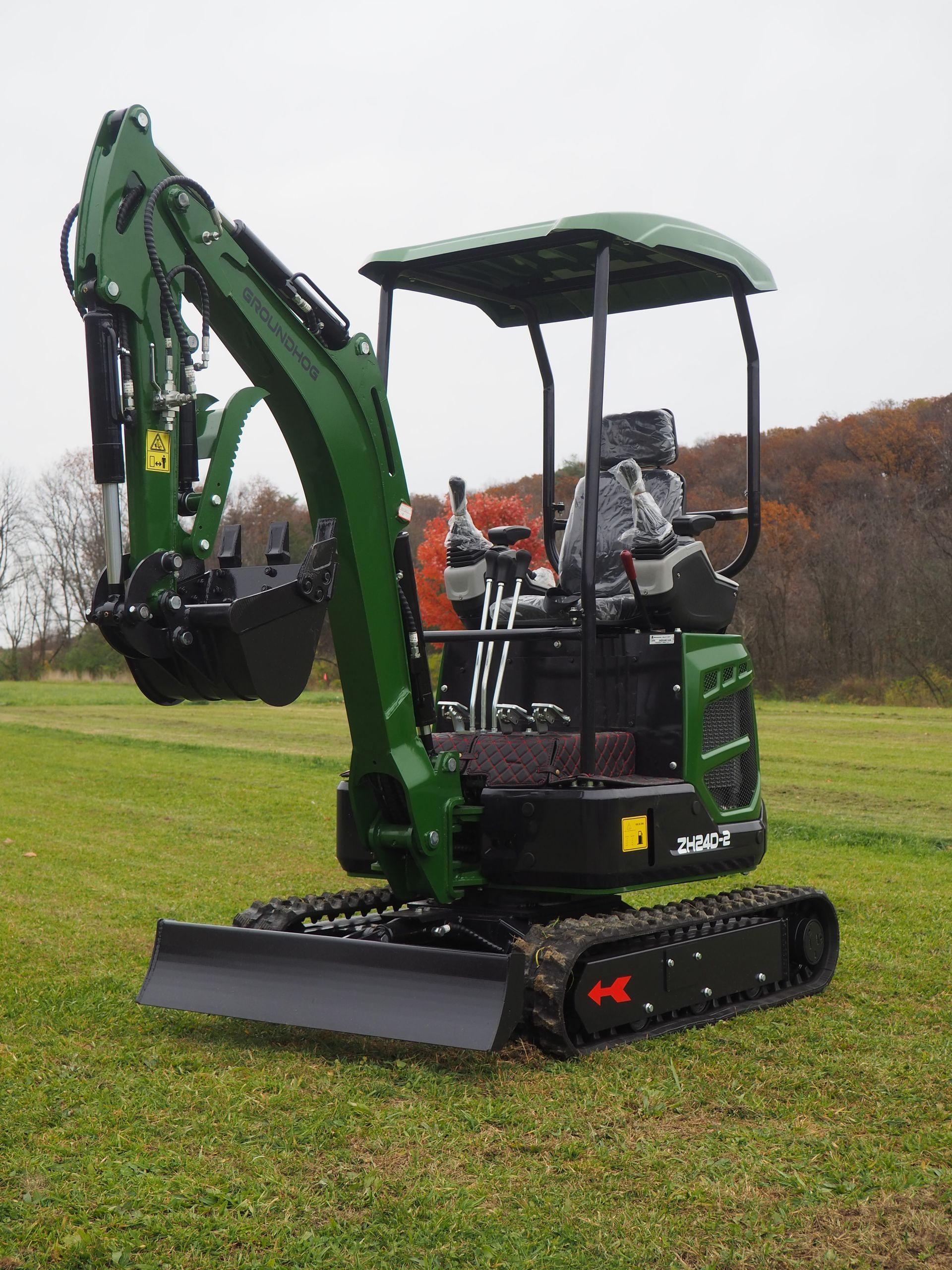 A green and black mini excavator stands in a grassy field with a canopy overhead and a blade on the front.