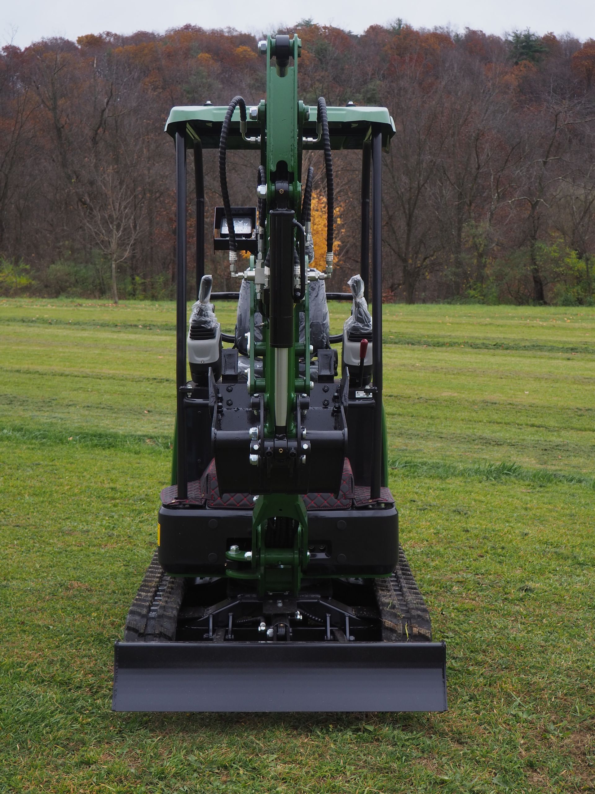 A green mini excavator parked in a field, viewed from the front, with its blade lowered to the grass.