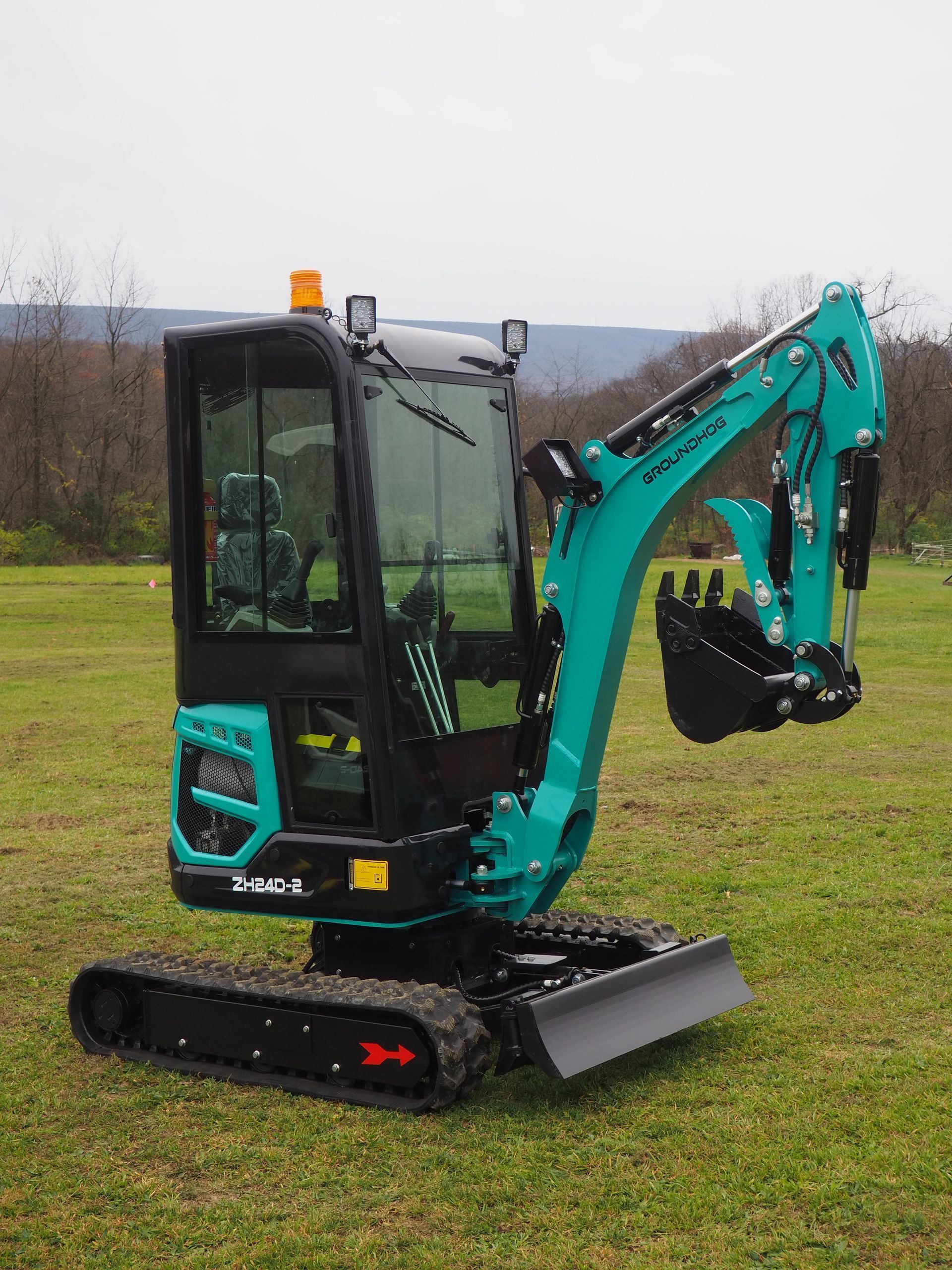A teal and black mini excavator sits on a grassy field under a cloudy sky.