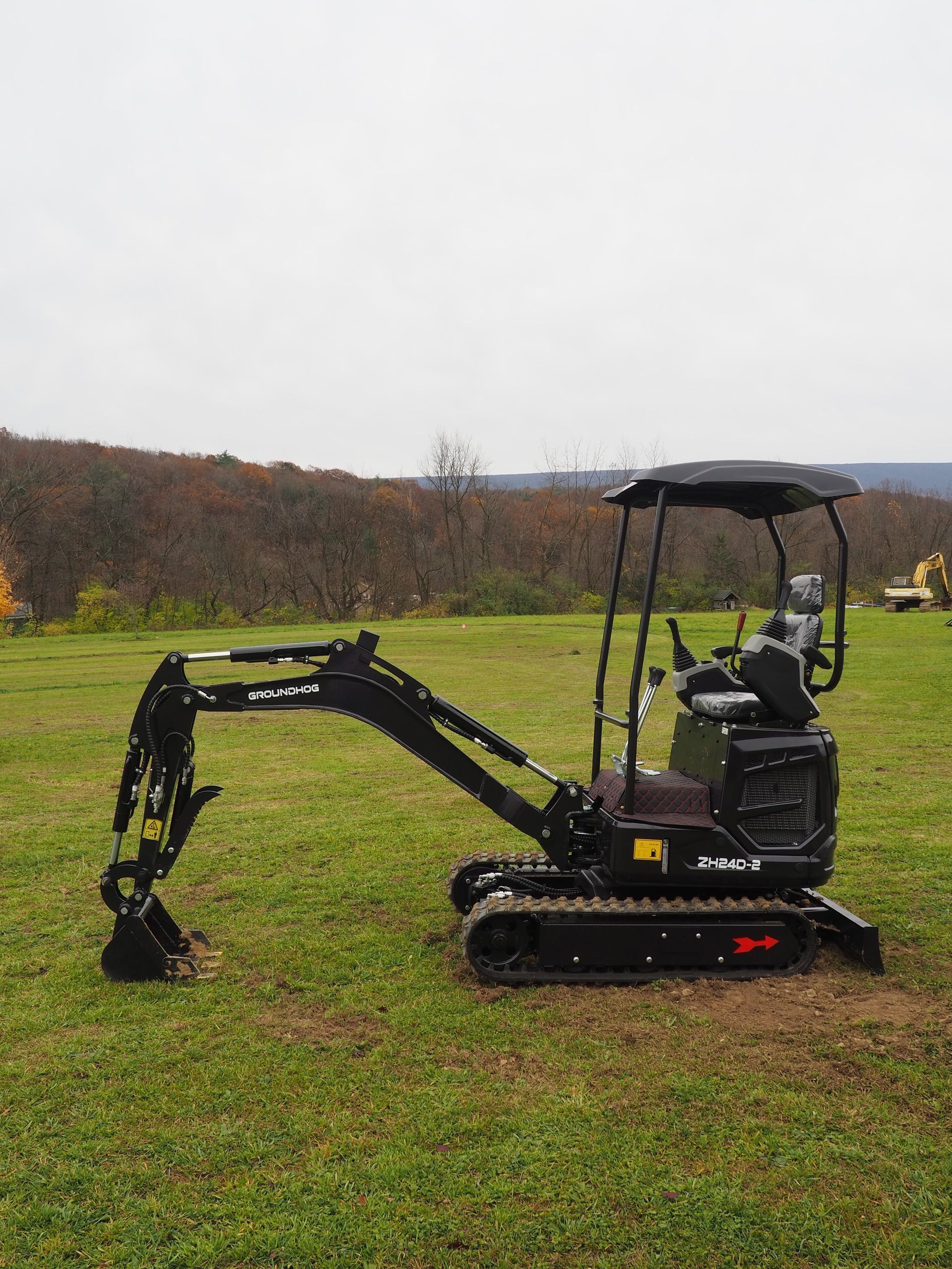 A small, black, tracked excavator sits in a grassy field on an overcast day with hills in the background.