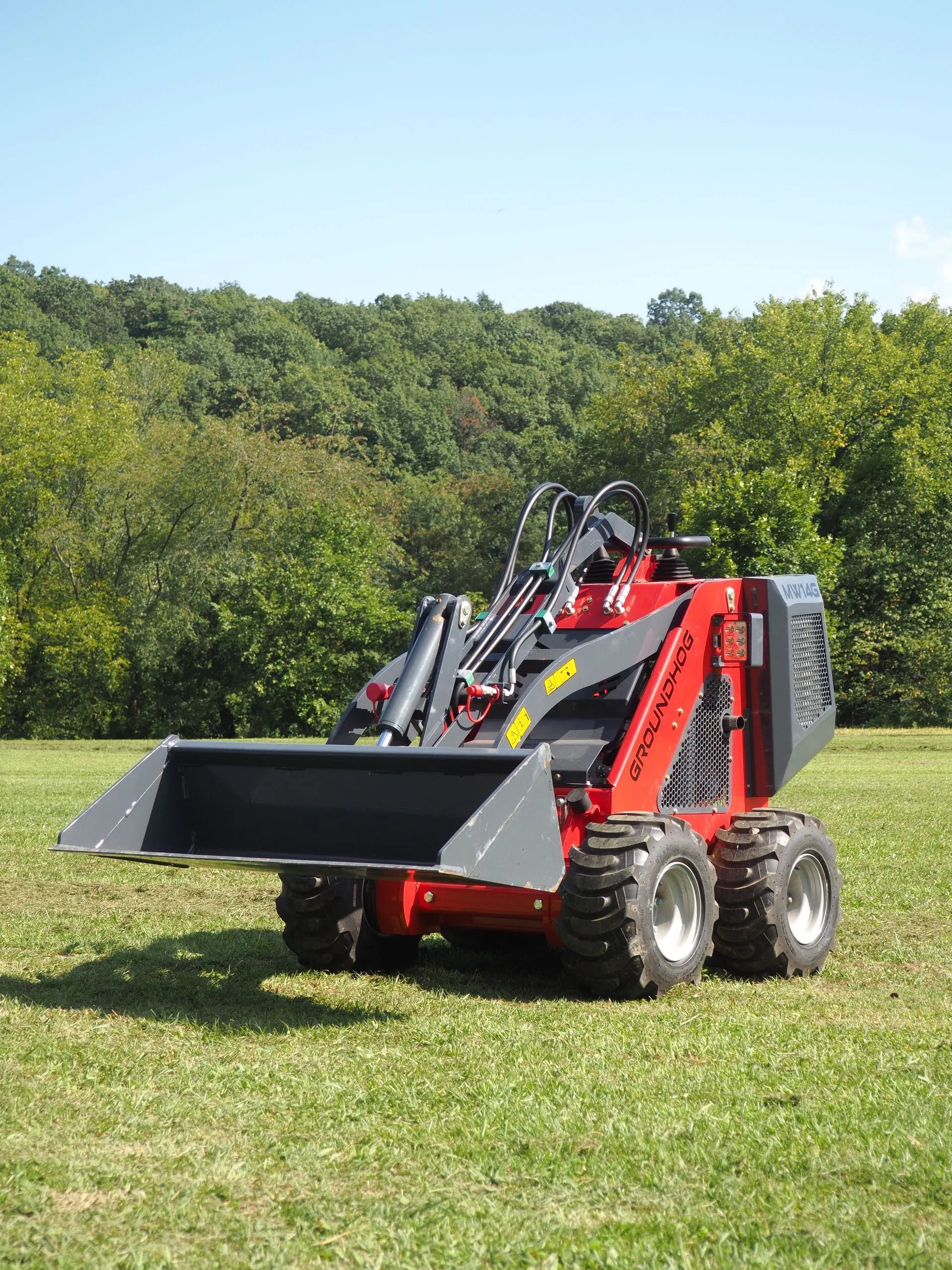 A red skid-steer loader with a black front bucket parked on a grassy field in front of a tree line.