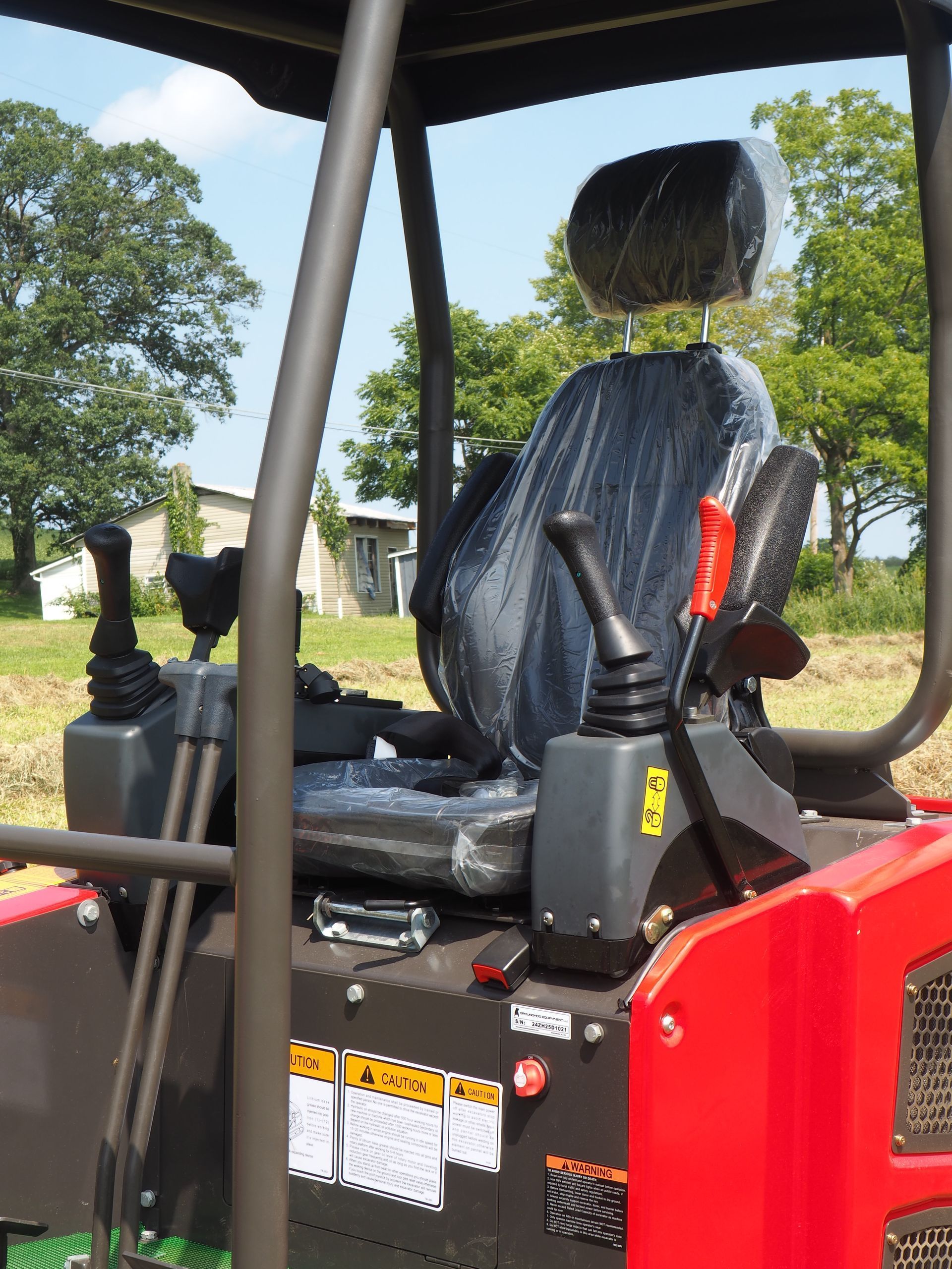 A red, open-cab industrial excavator seat with black controls, protected by plastic film, outdoors in a grassy field.