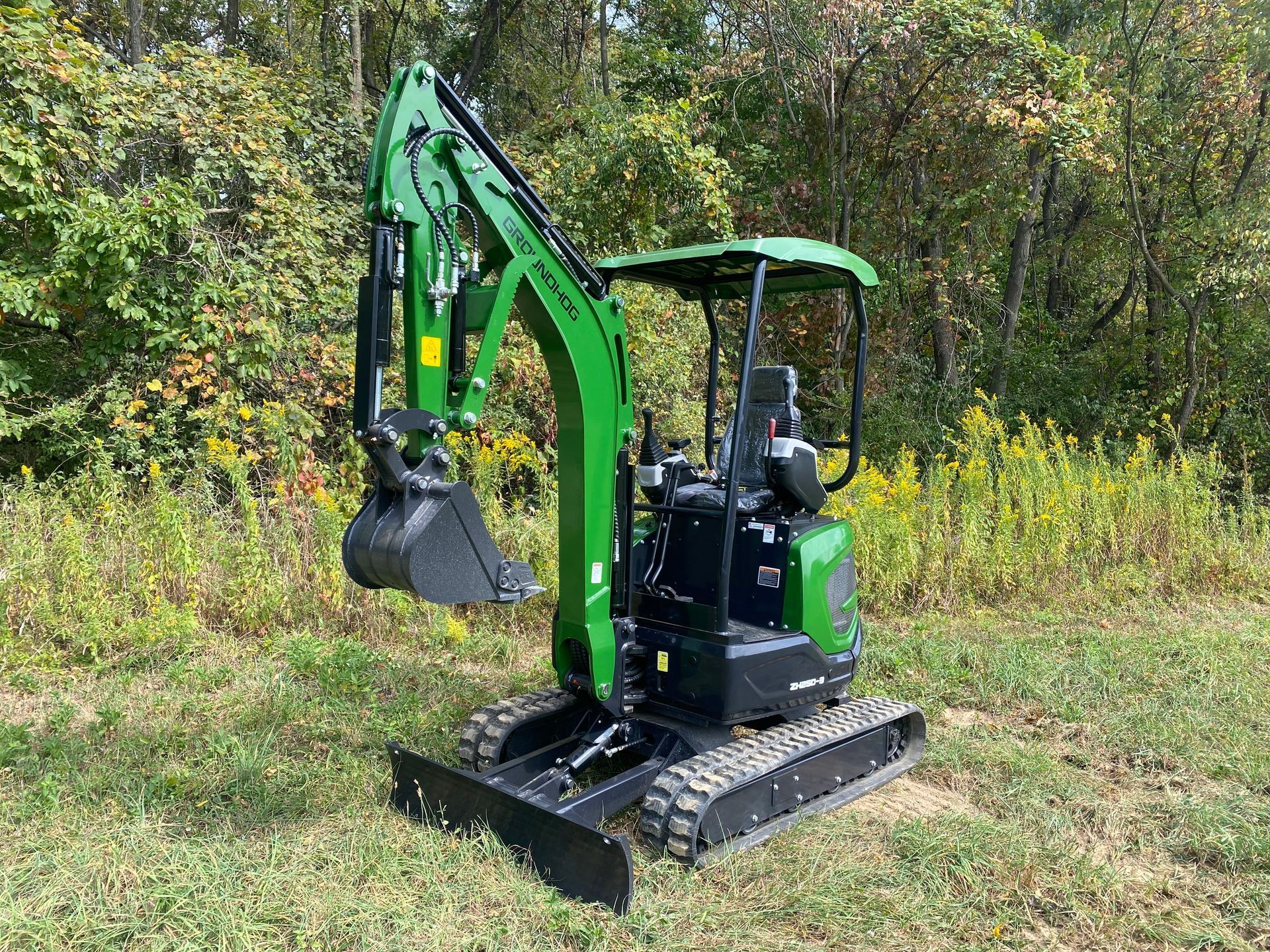 A green and black compact excavator sits on a grassy field in front of a line of trees.