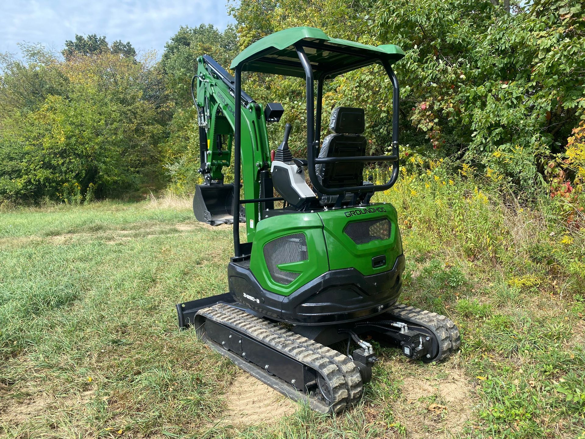 A green mini excavator parked on a grassy field with trees in the background.