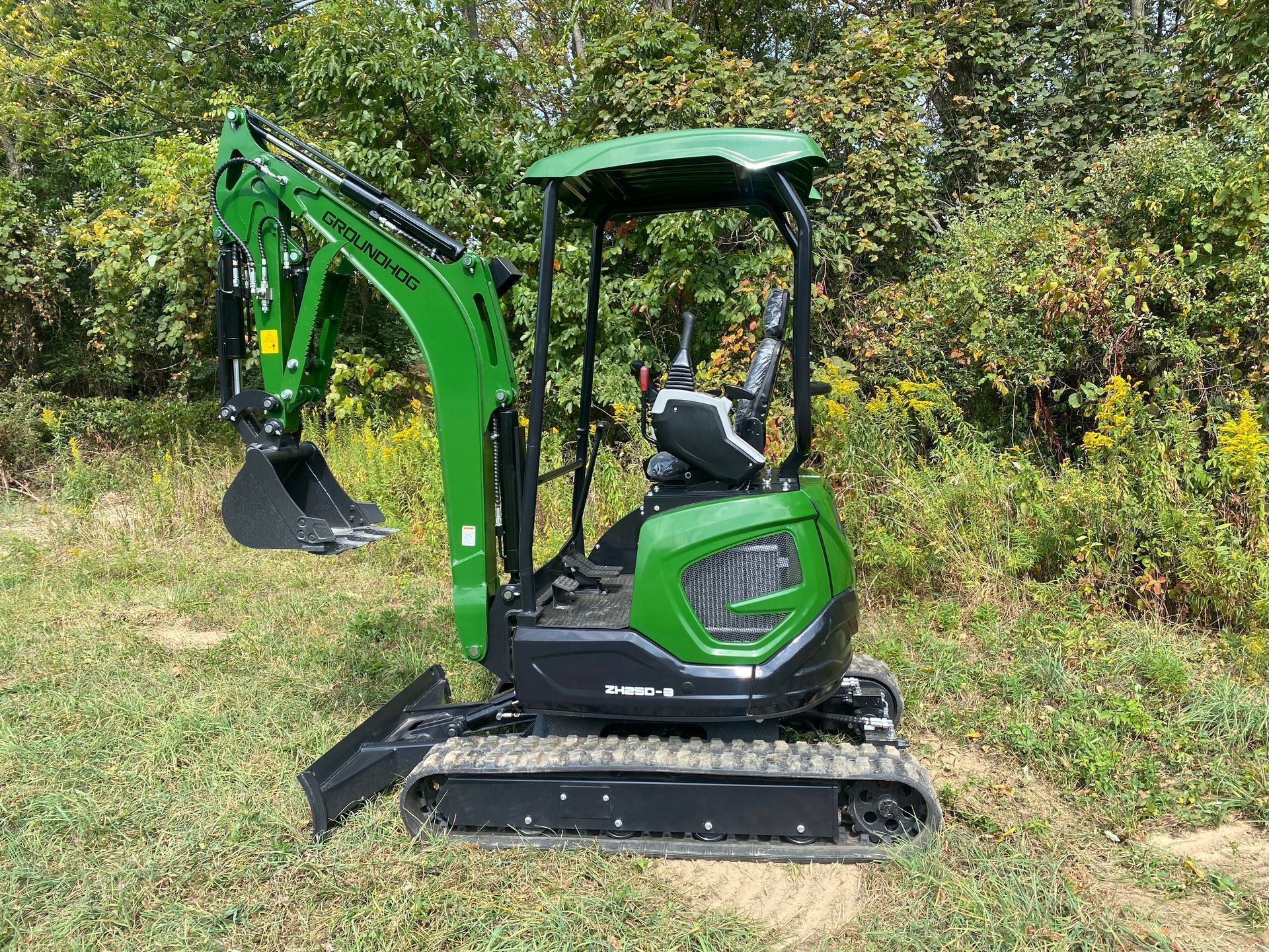 A compact green excavator with a black bucket and tracks parked on a grassy field in front of a tree line.