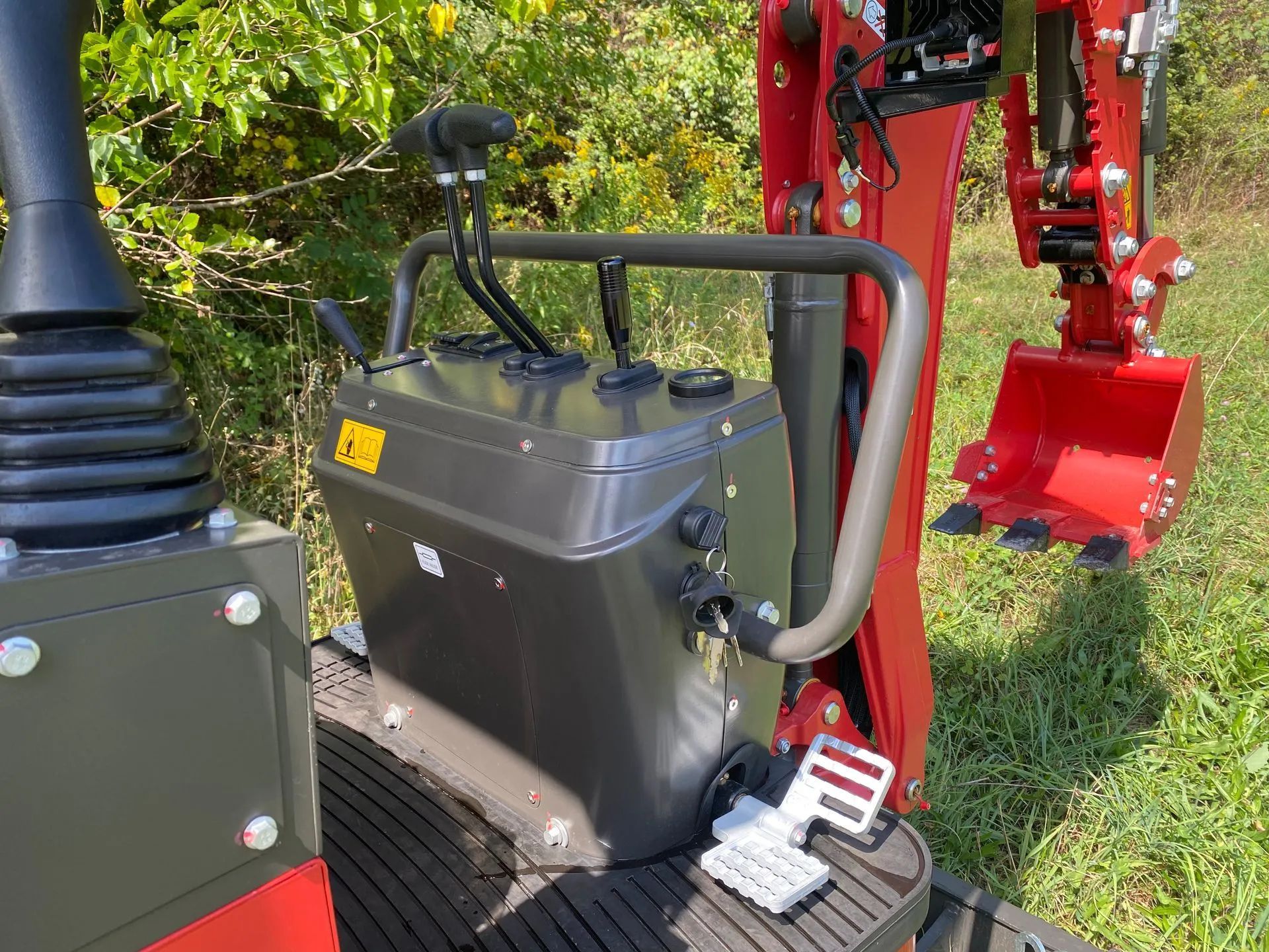 A close-up of a mini excavator's control console and metal safety bar, with the red digging arm and bucket in the back.
