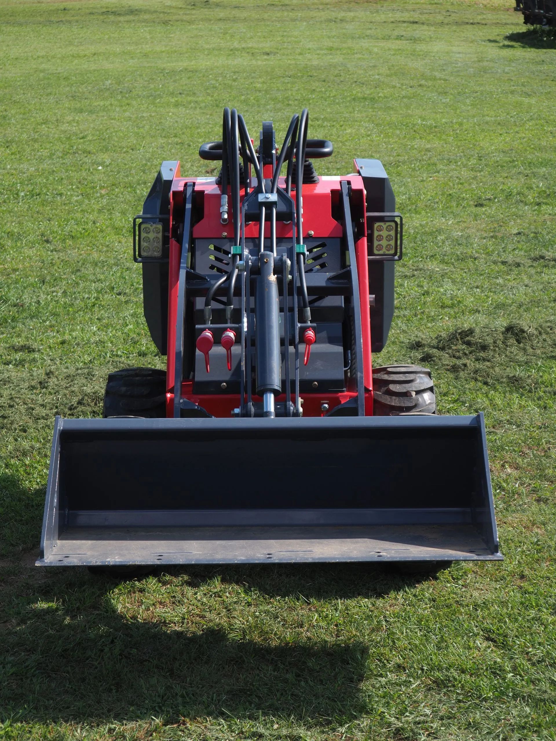 A red mini skid steer loader with a metal bucket sits on a grassy field.