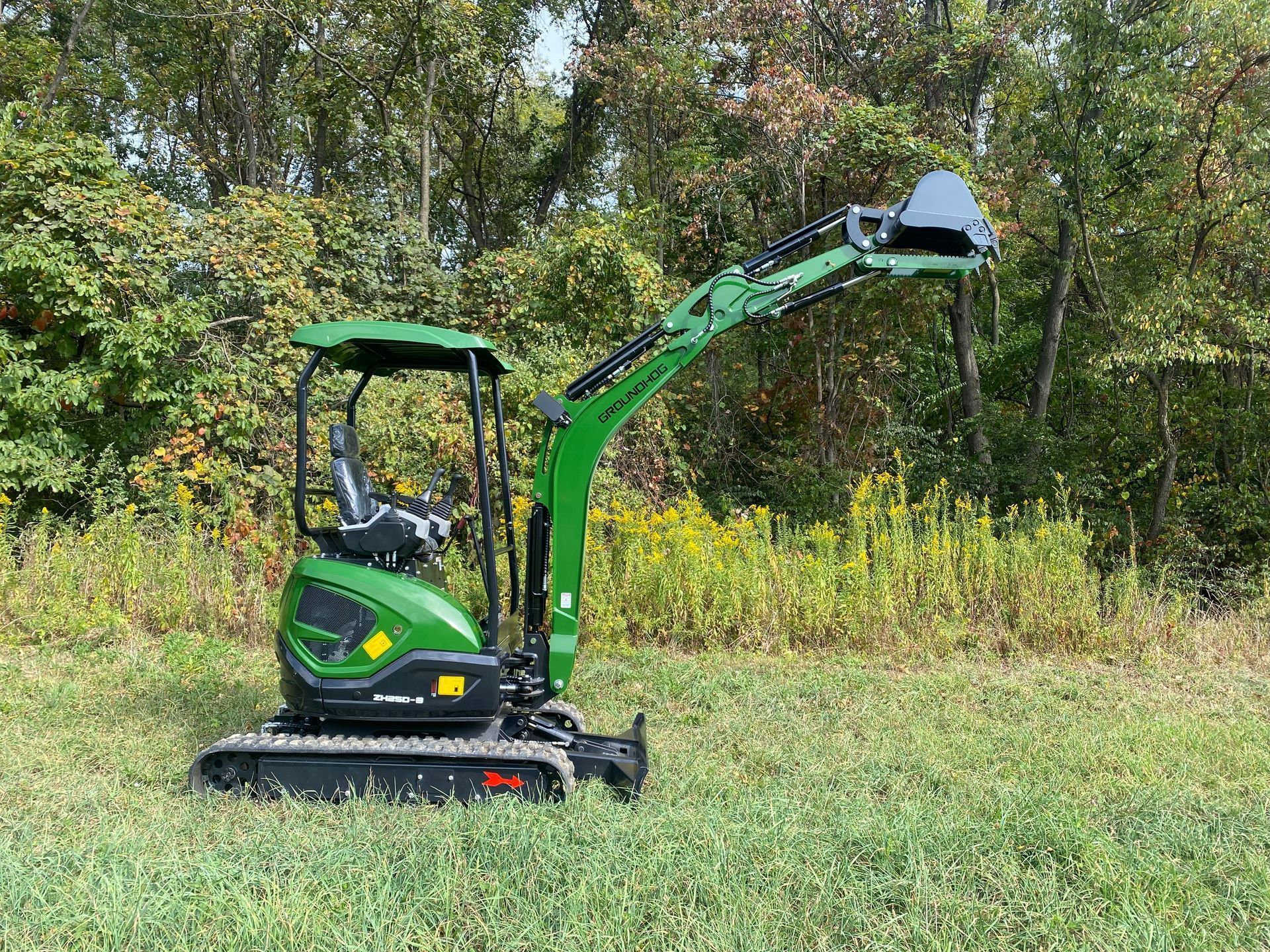 A green John Deere mini excavator parked in a grassy field with trees in the background.