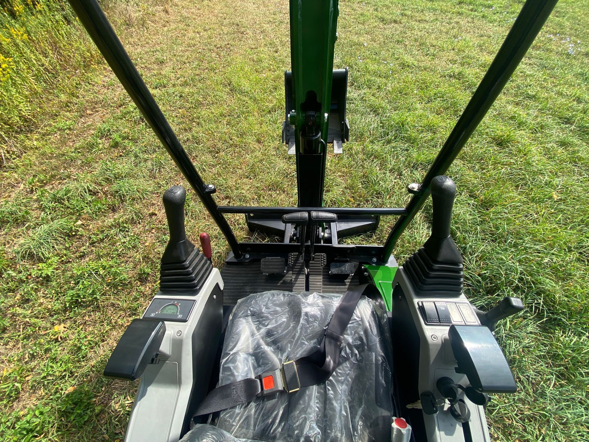 View from the driver's seat of an excavator, showing joysticks, a covered seat, and the green boom in a grassy field.