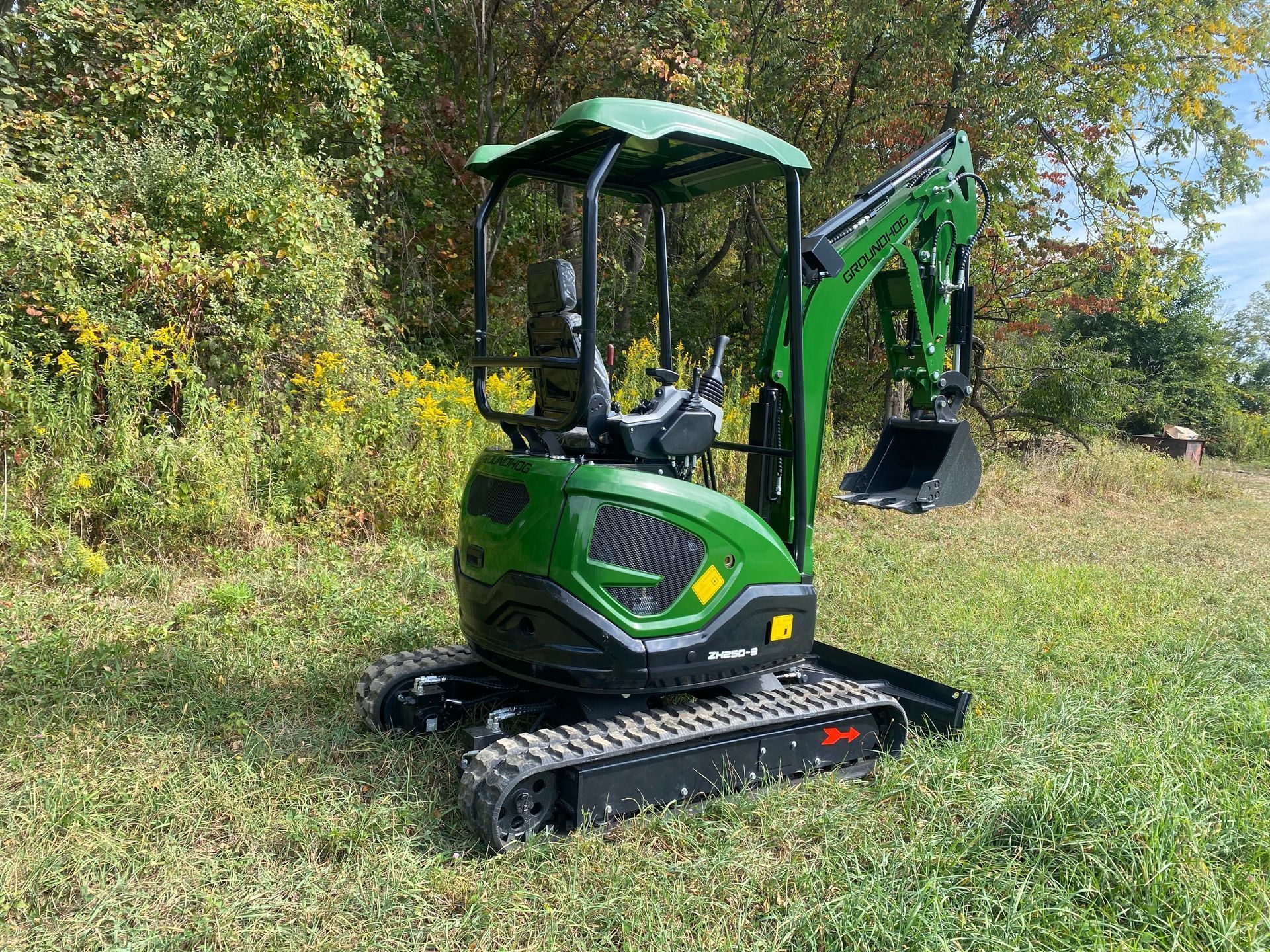 A green John Deere compact excavator parked in a grassy, wooded area.