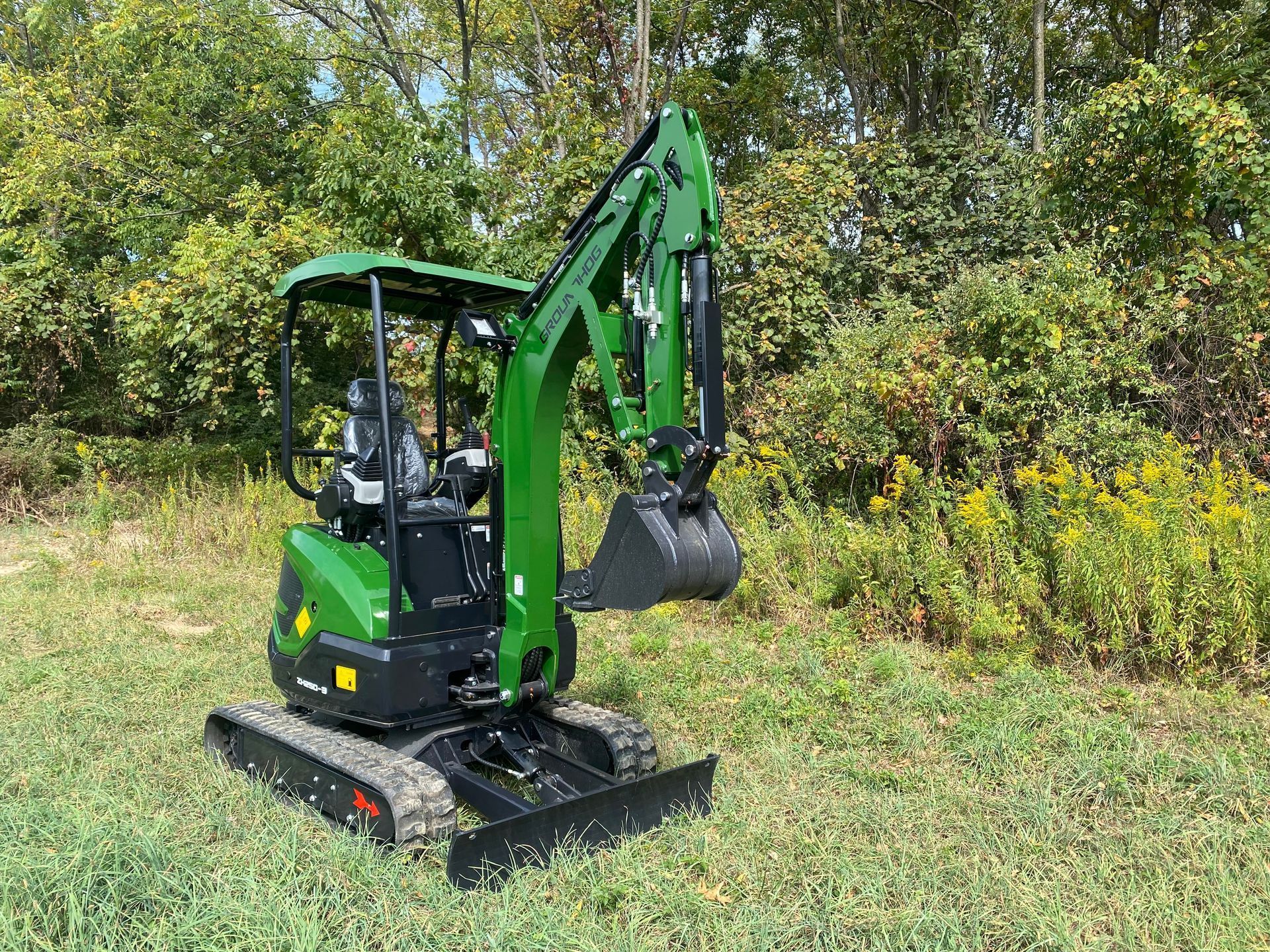 A compact green mini excavator parked on a grassy field in front of a tree line.
