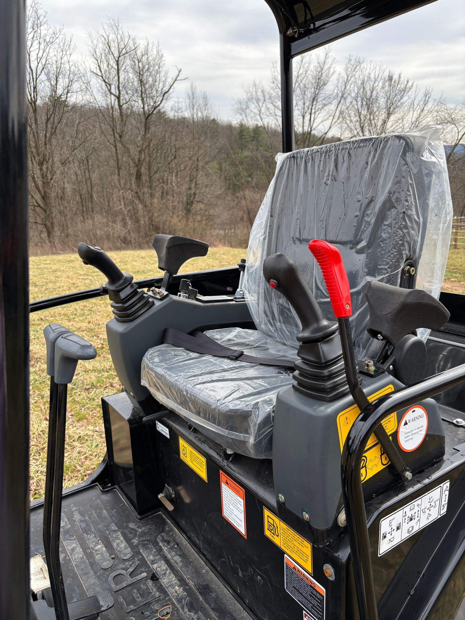 The operator station of a compact excavator featuring a seat, control joysticks, and safety levers outdoors.