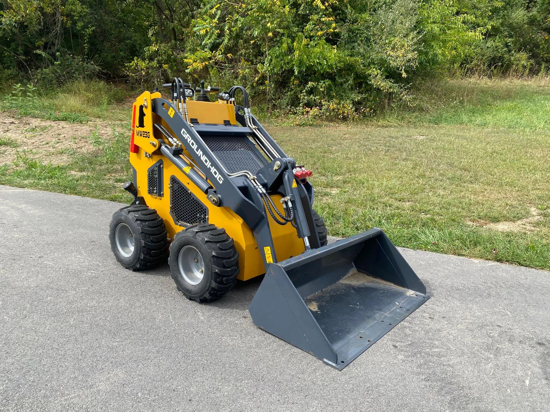 A yellow compact skid steer loader with a gray bucket sits on an asphalt path against a background of trees.