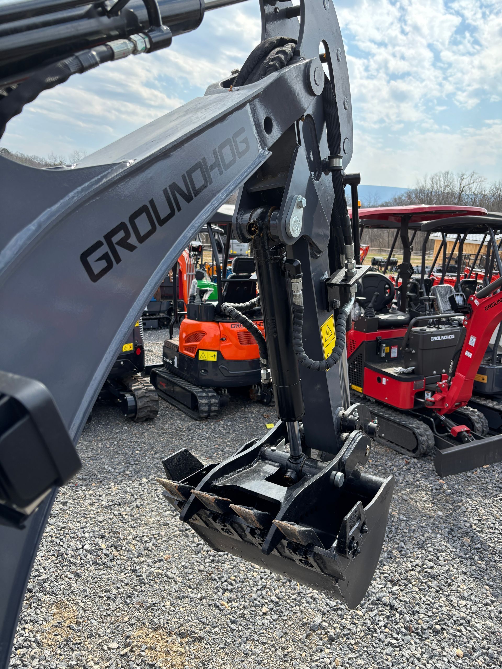 A gray GroundHog mini-excavator arm with a bucket attached, parked in a lot with other construction equipment.