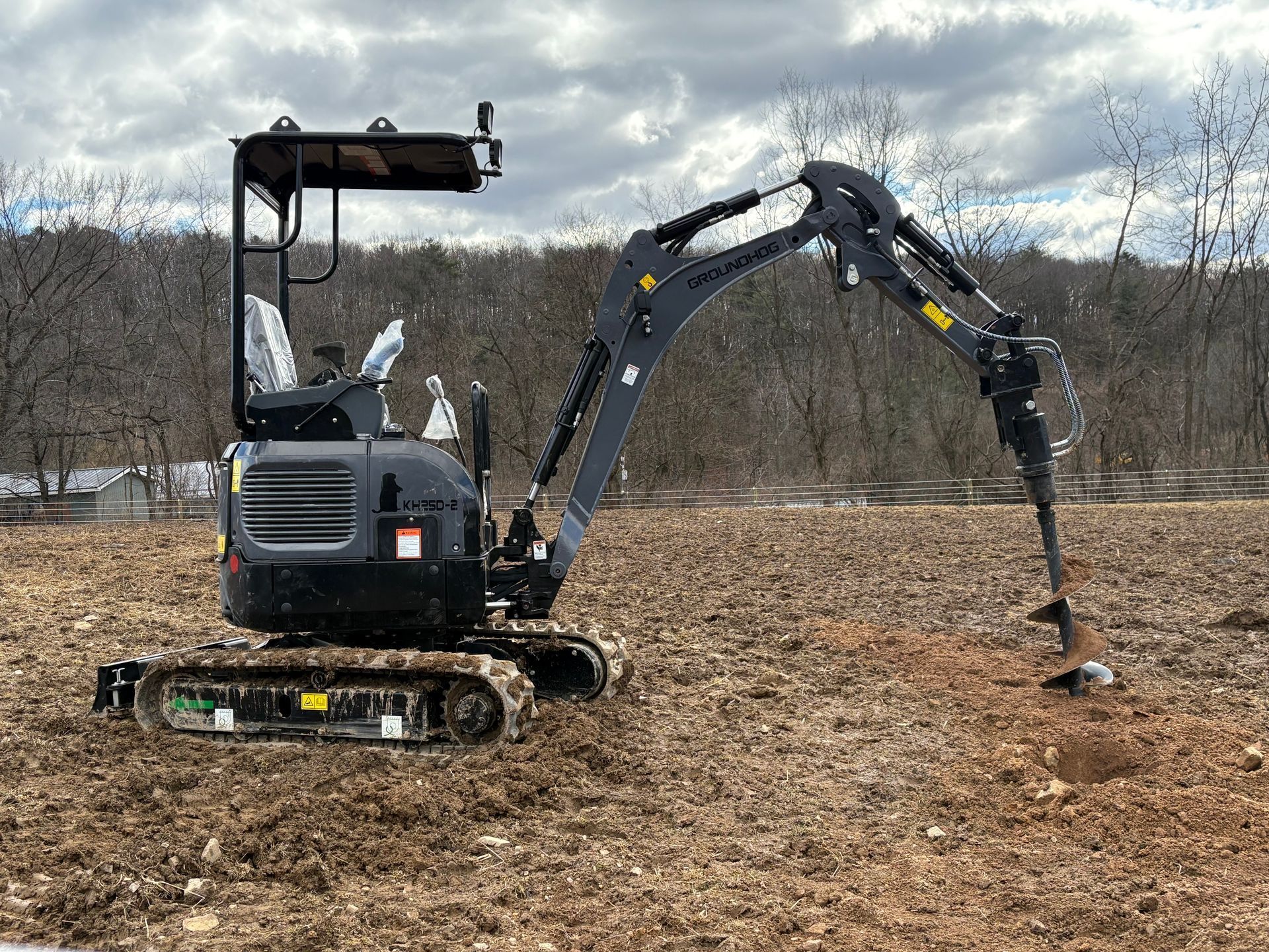 A gray compact excavator with an auger attachment drills into brown, tilled soil in an open field.