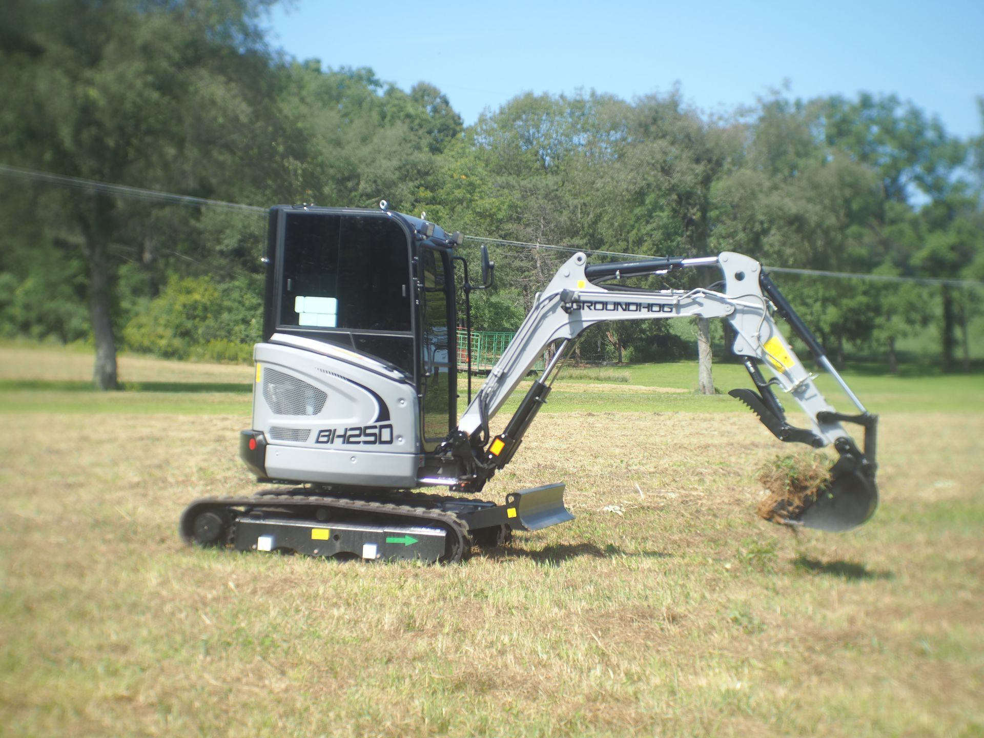 A silver and black compact excavator sits in a grassy field, its bucket raised above the ground.