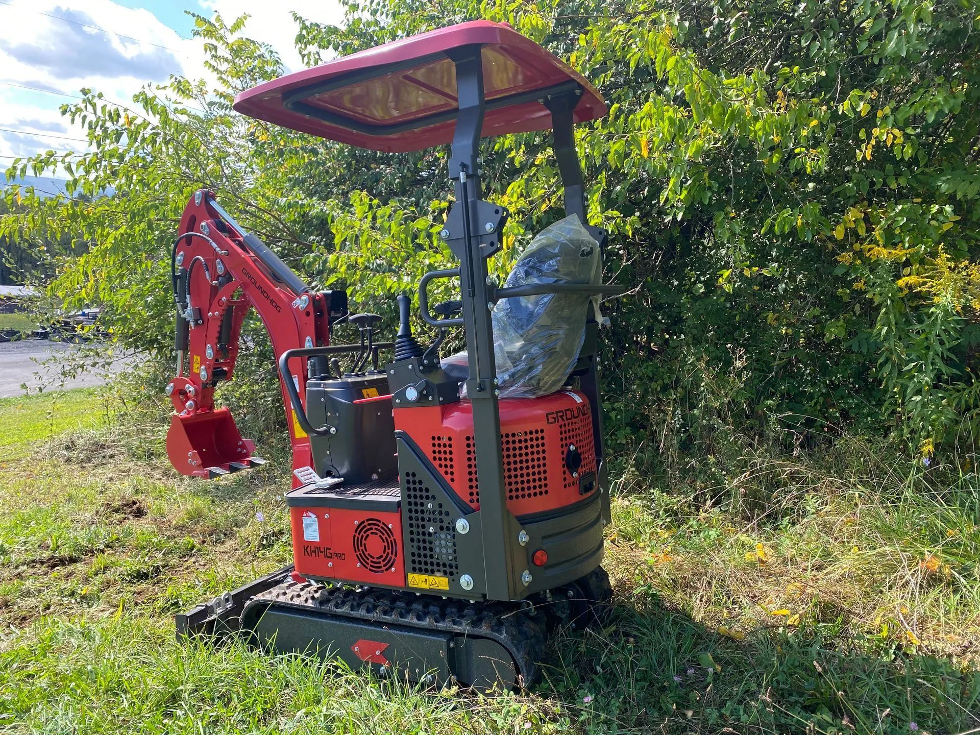 A red compact excavator parked on a grassy area in front of green trees.