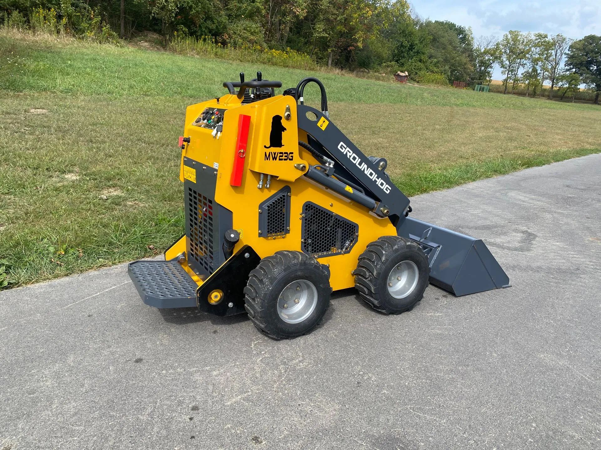 A yellow and black stand-on mini skid steer loader parked on a paved path next to a grassy hill.