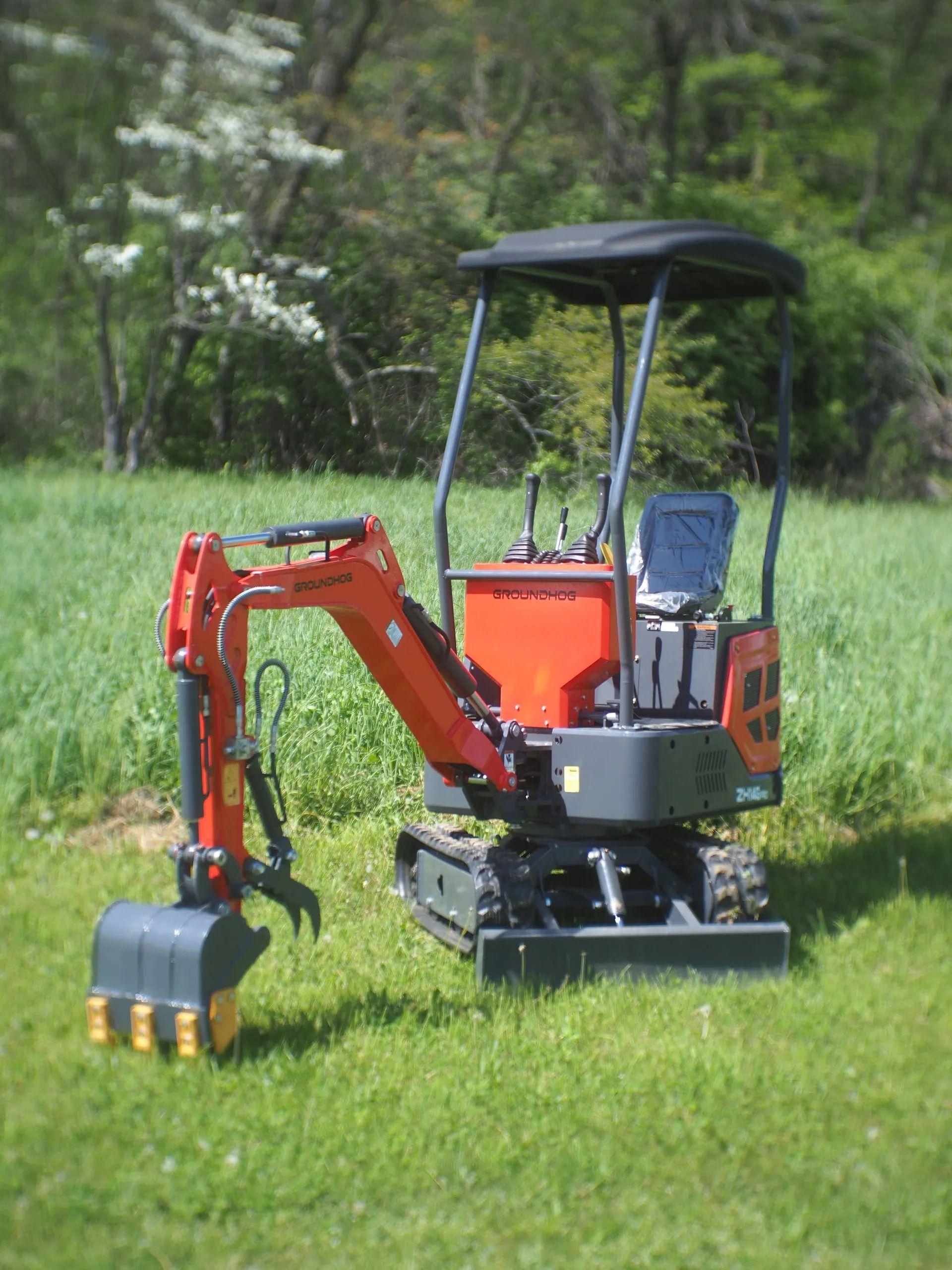A bright orange and gray mini excavator parked on a grassy field with a tree-lined background.