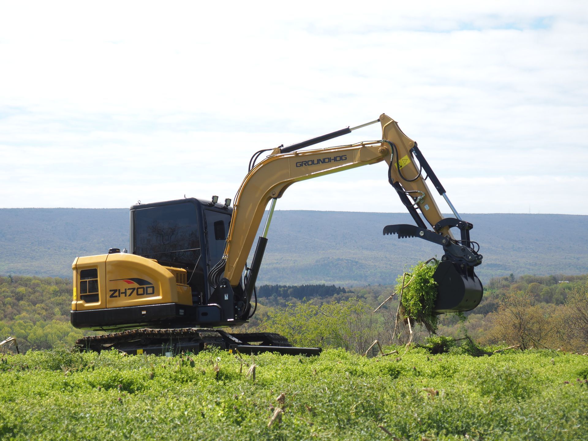 A yellow excavator with a bucket attachment clearing brush in a grassy field with rolling hills in the background.