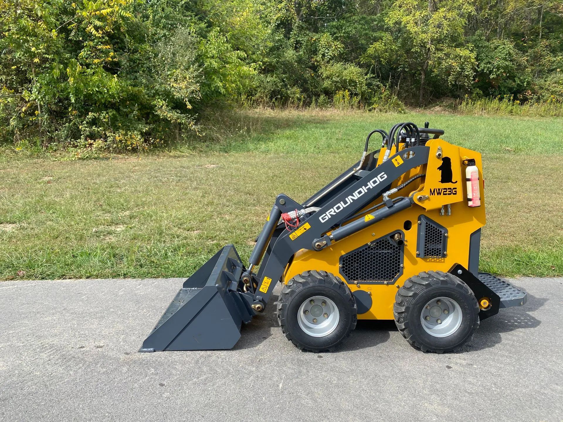 A yellow and grey GroundHog skid steer loader parked on a paved path in front of a grassy, wooded area.