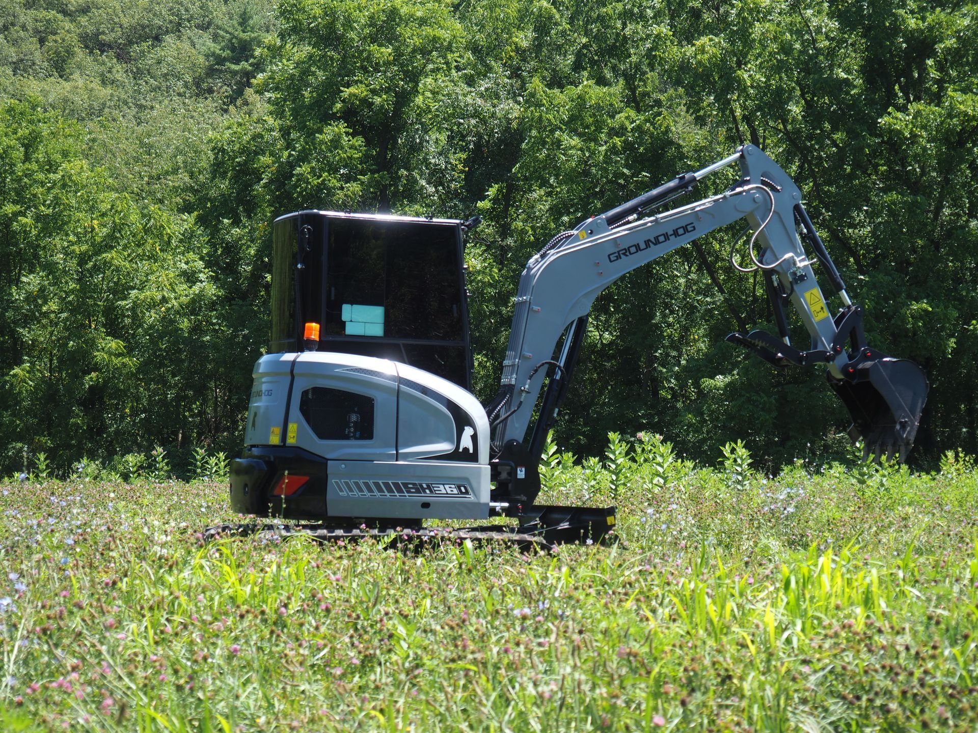 A grey mini-excavator sits parked in a lush green field with a dense treeline in the background.