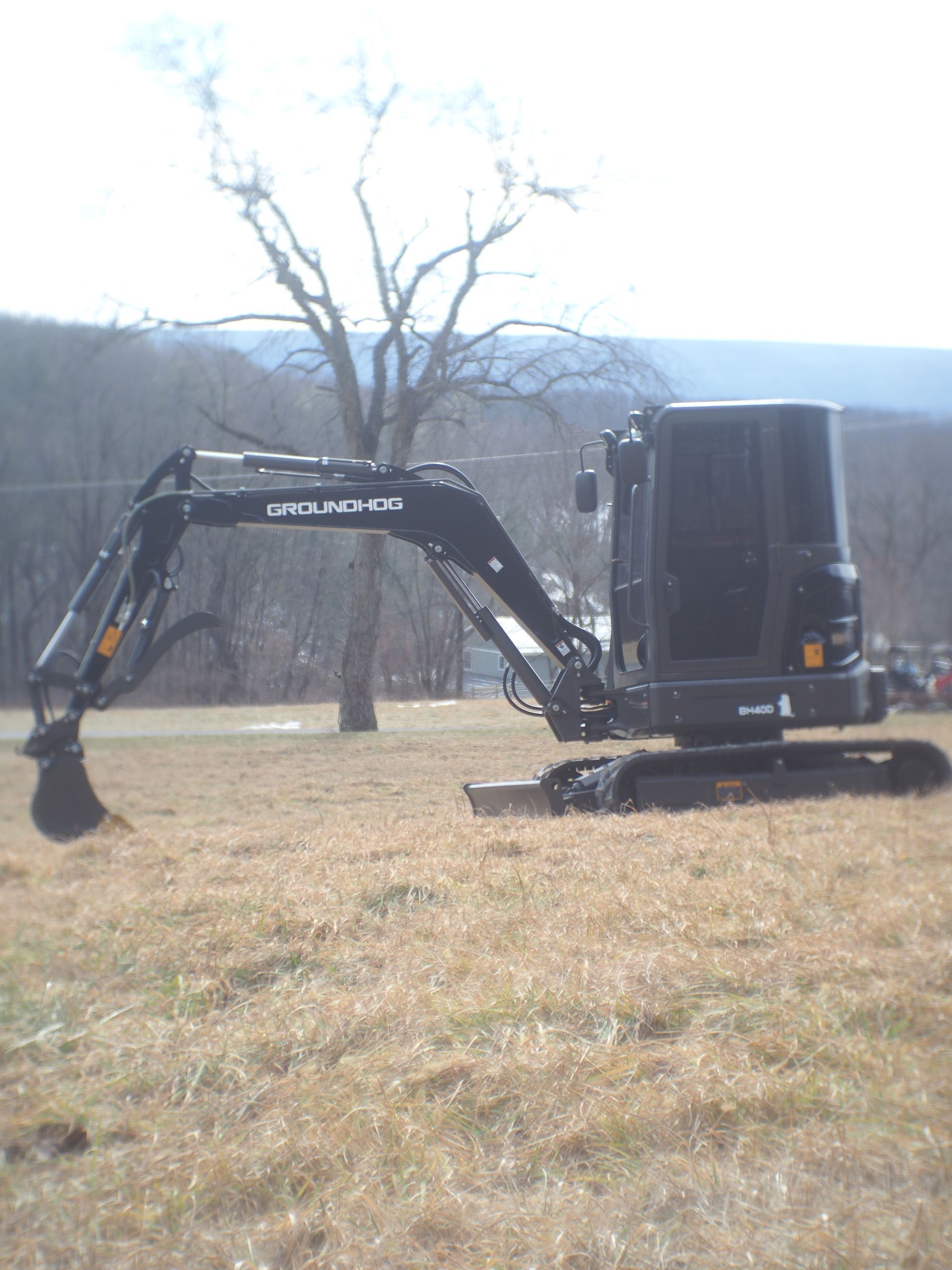 A black mini excavator parked on a grassy field in front of a tree and distant hills.