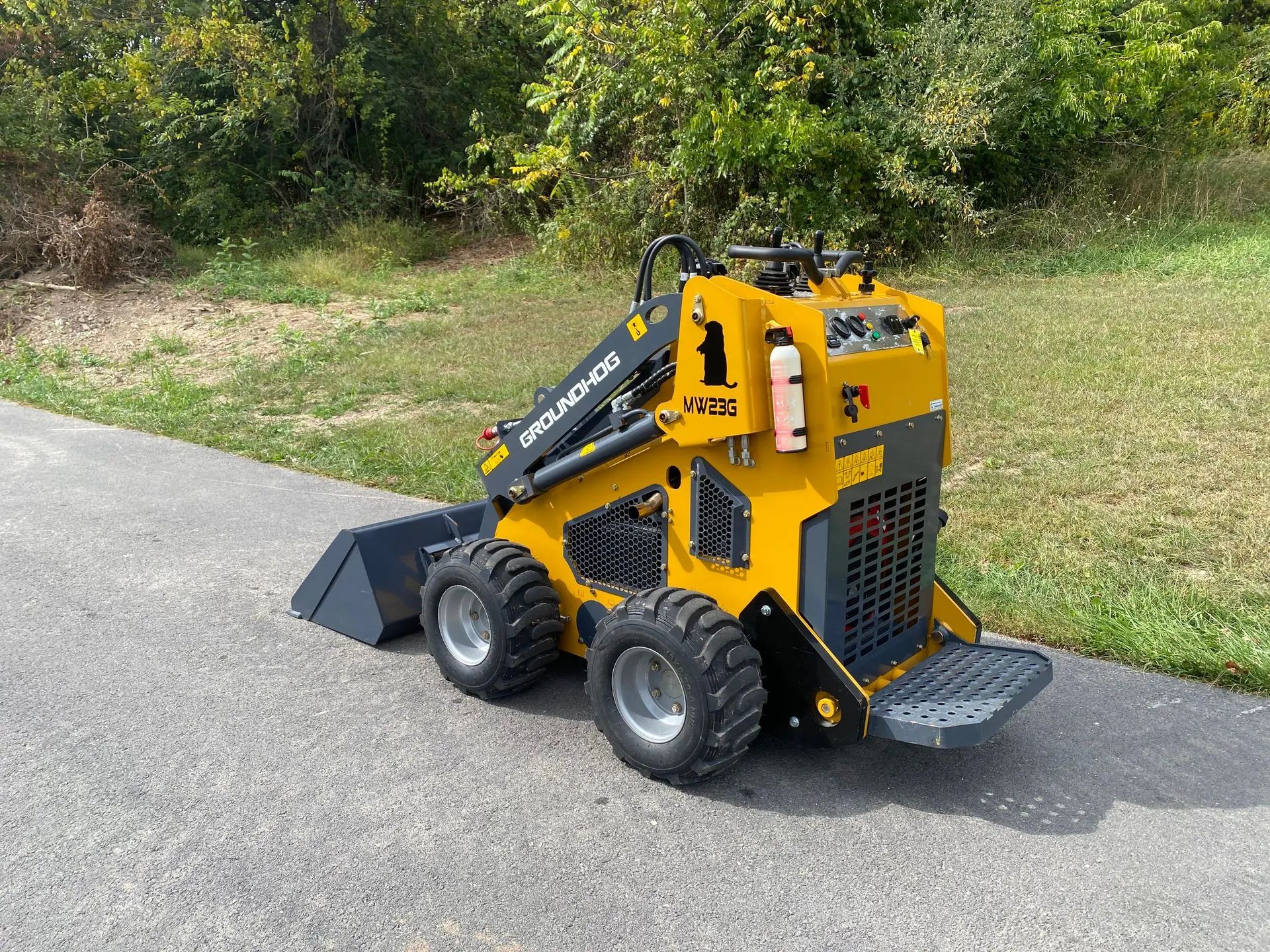 A yellow stand-on mini skid steer loader with a bucket attachment sits on an asphalt path next to a grassy area.
