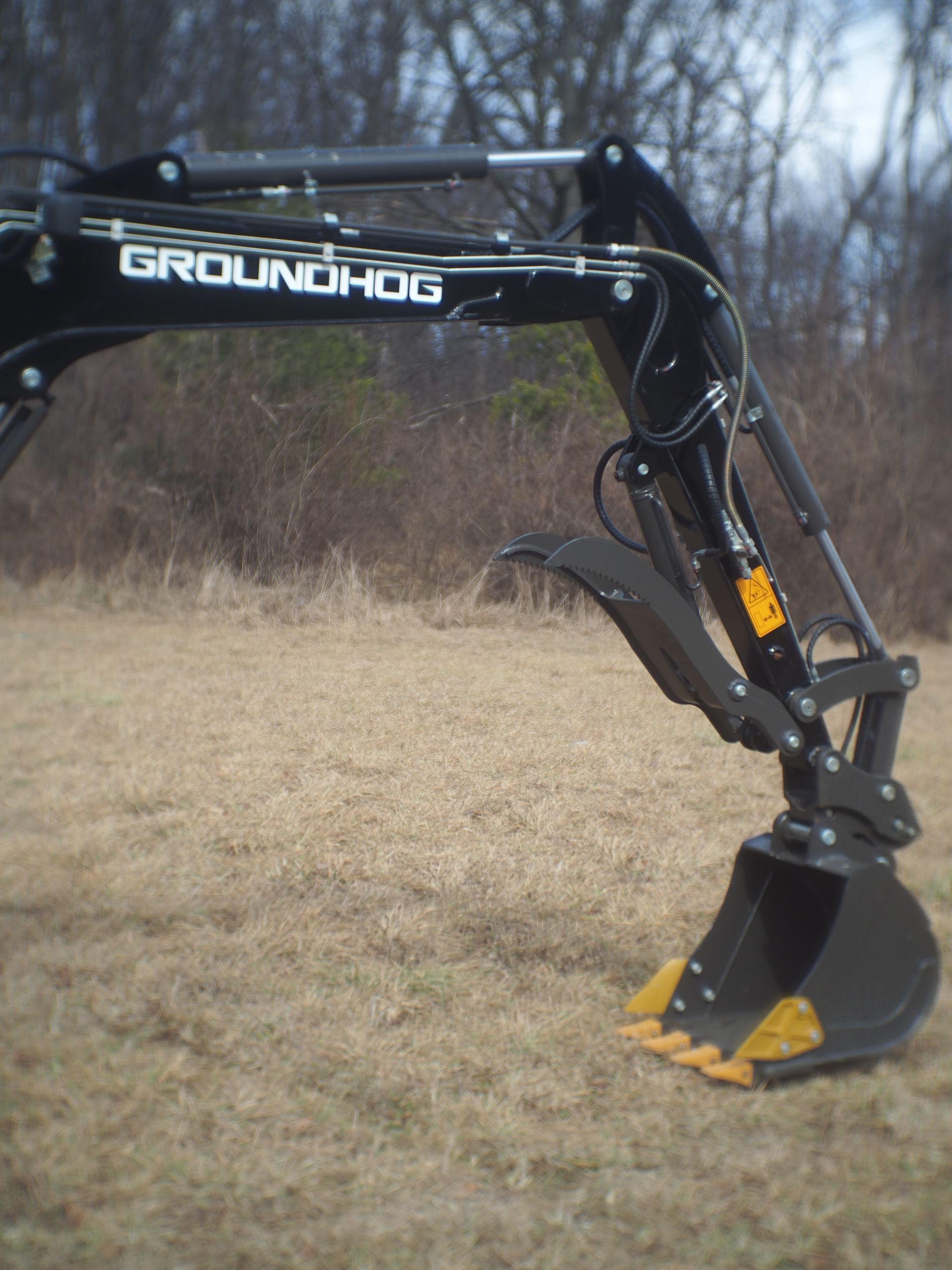 A black GroundHog excavator arm with a bucket and hydraulic grapple, positioned against an outdoor grassy background.