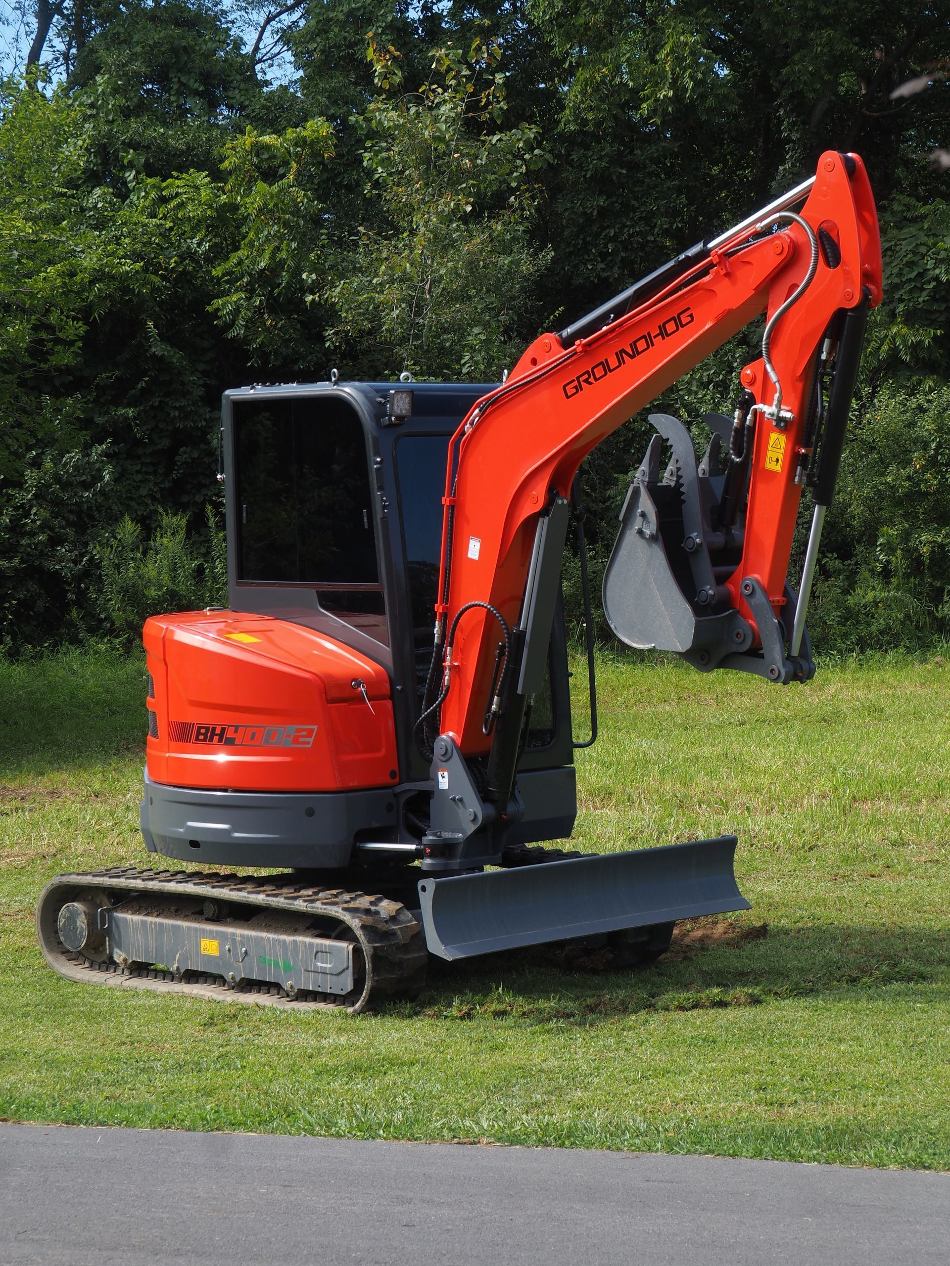 An orange compact excavator sits on a grassy field with a backdrop of trees.