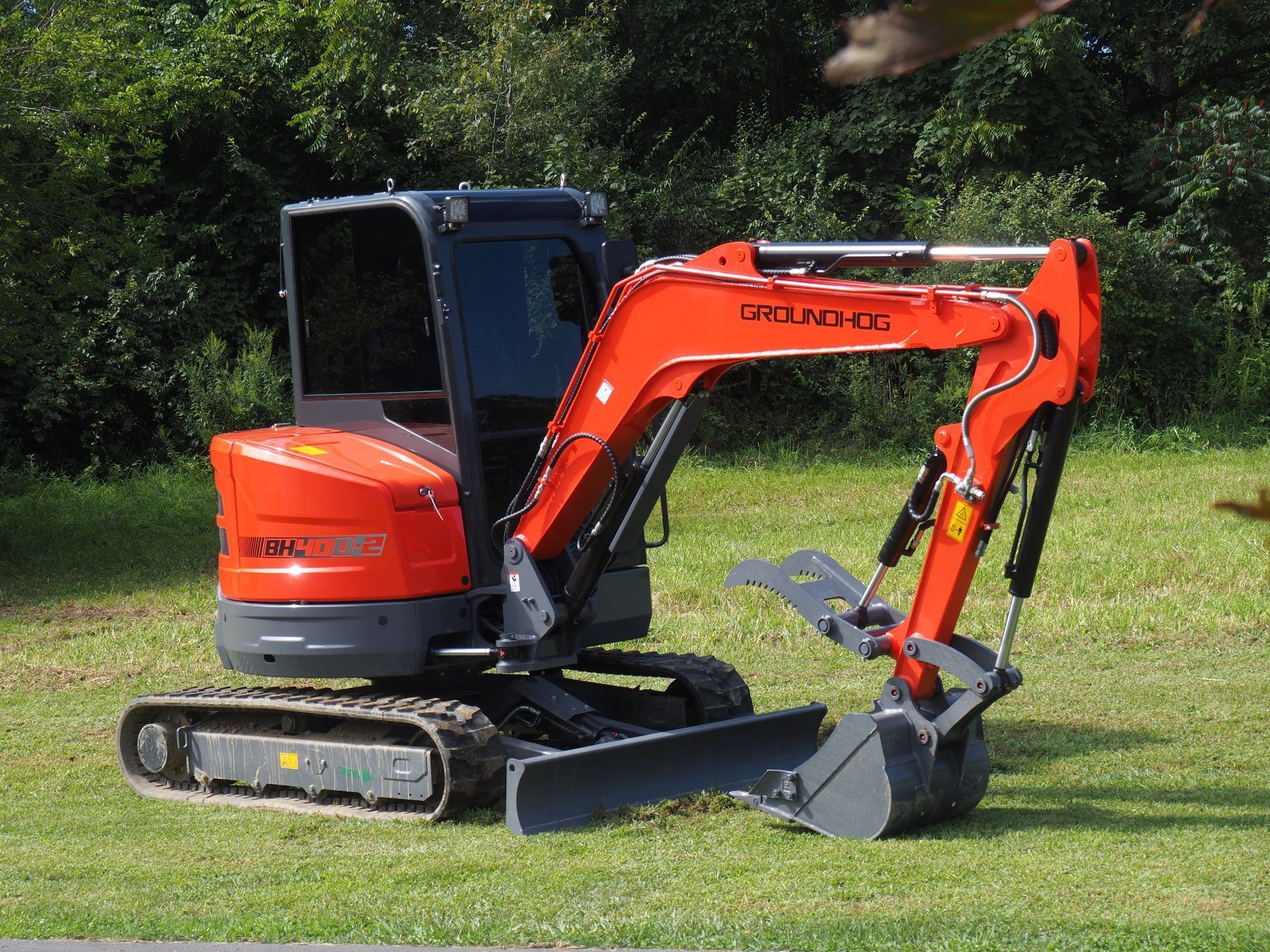 An orange and grey compact excavator parked on a grass field in front of a line of trees.