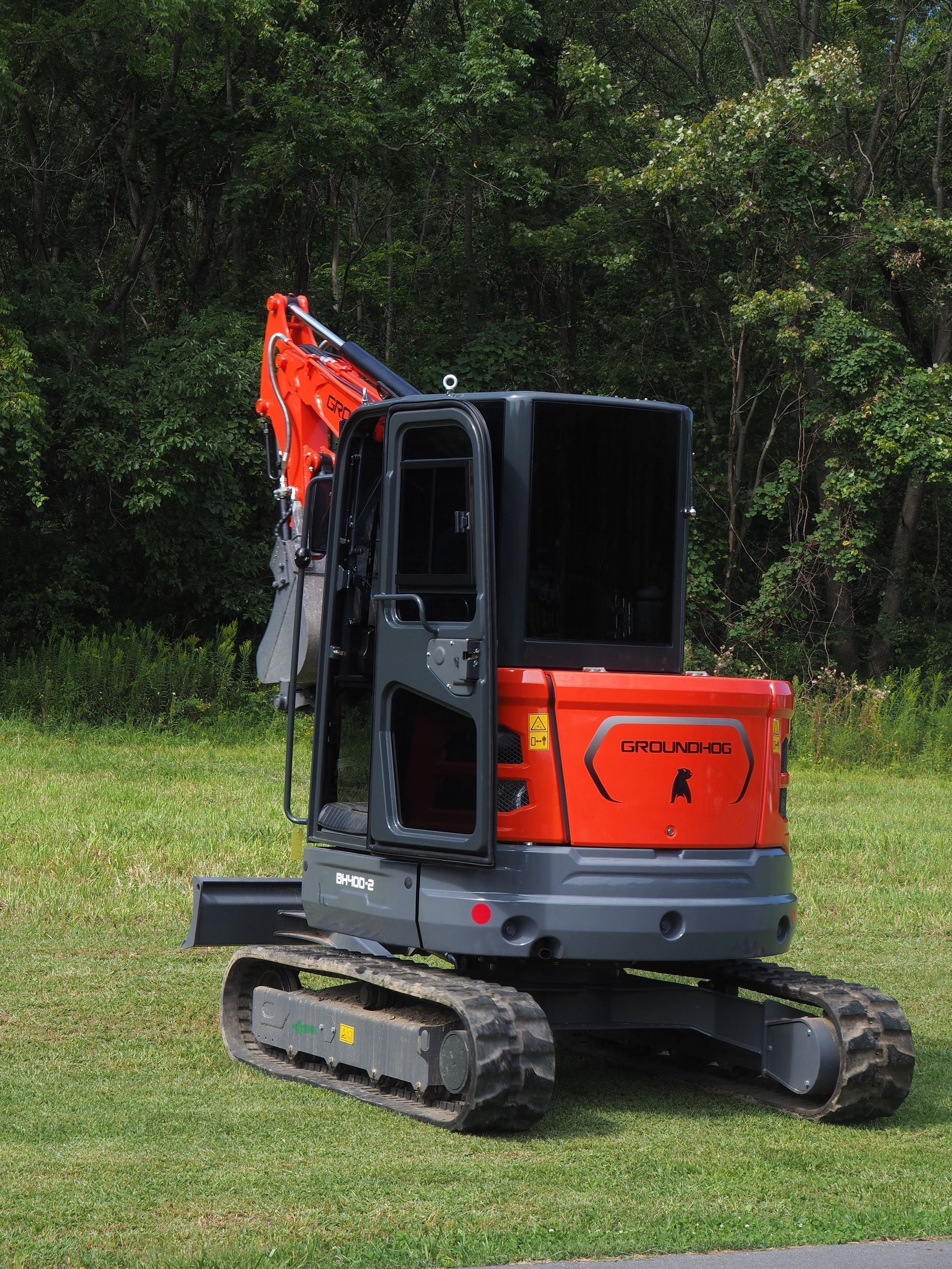 An orange and grey compact excavator parked on a grassy field in front of a tree line.