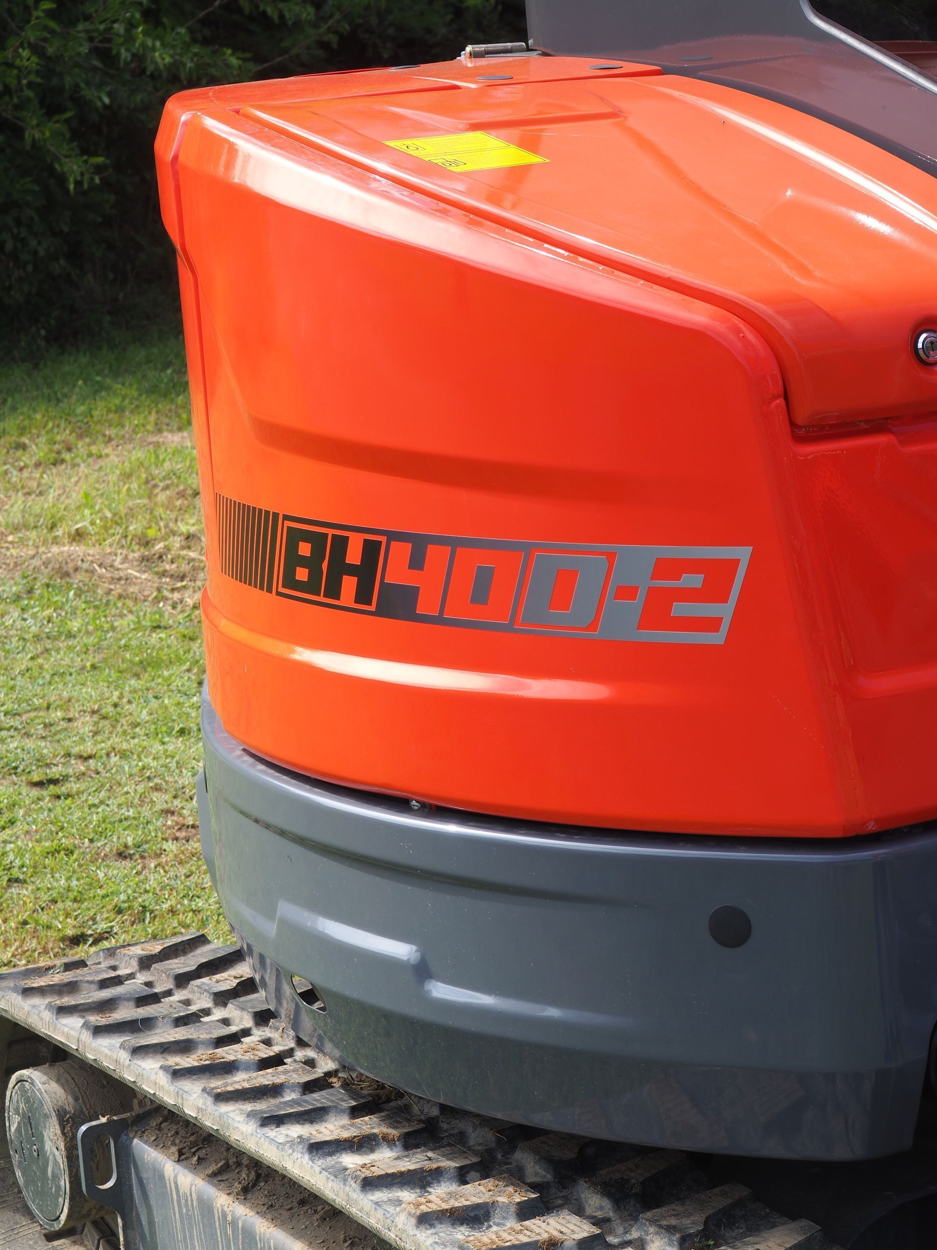 Orange rear body of a BH400-2 mini excavator with grey bumper and black crawler tracks, parked on a grassy field.