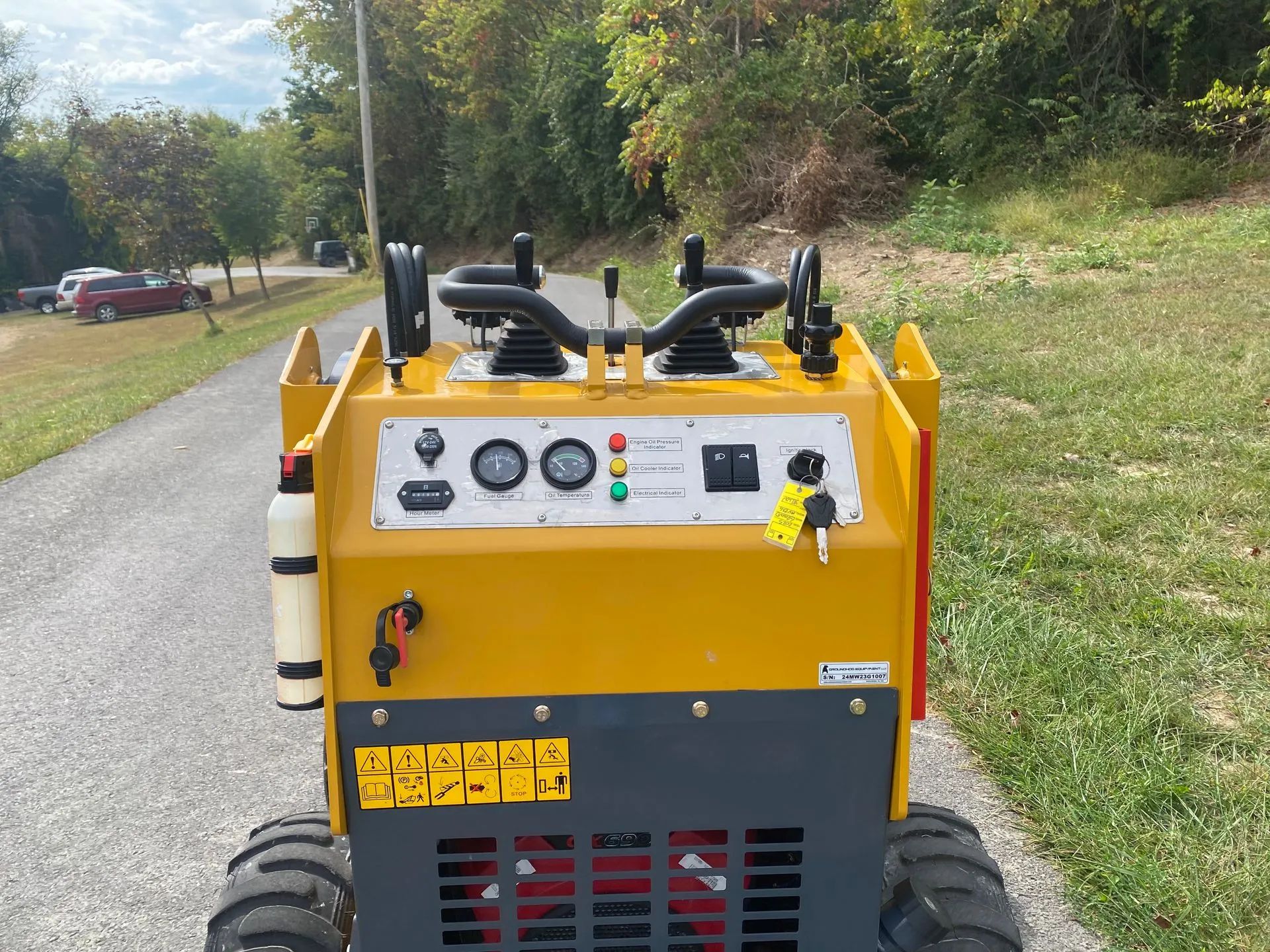 A bright yellow walk-behind mini skid steer loader on a paved path, viewed from behind the operator controls.