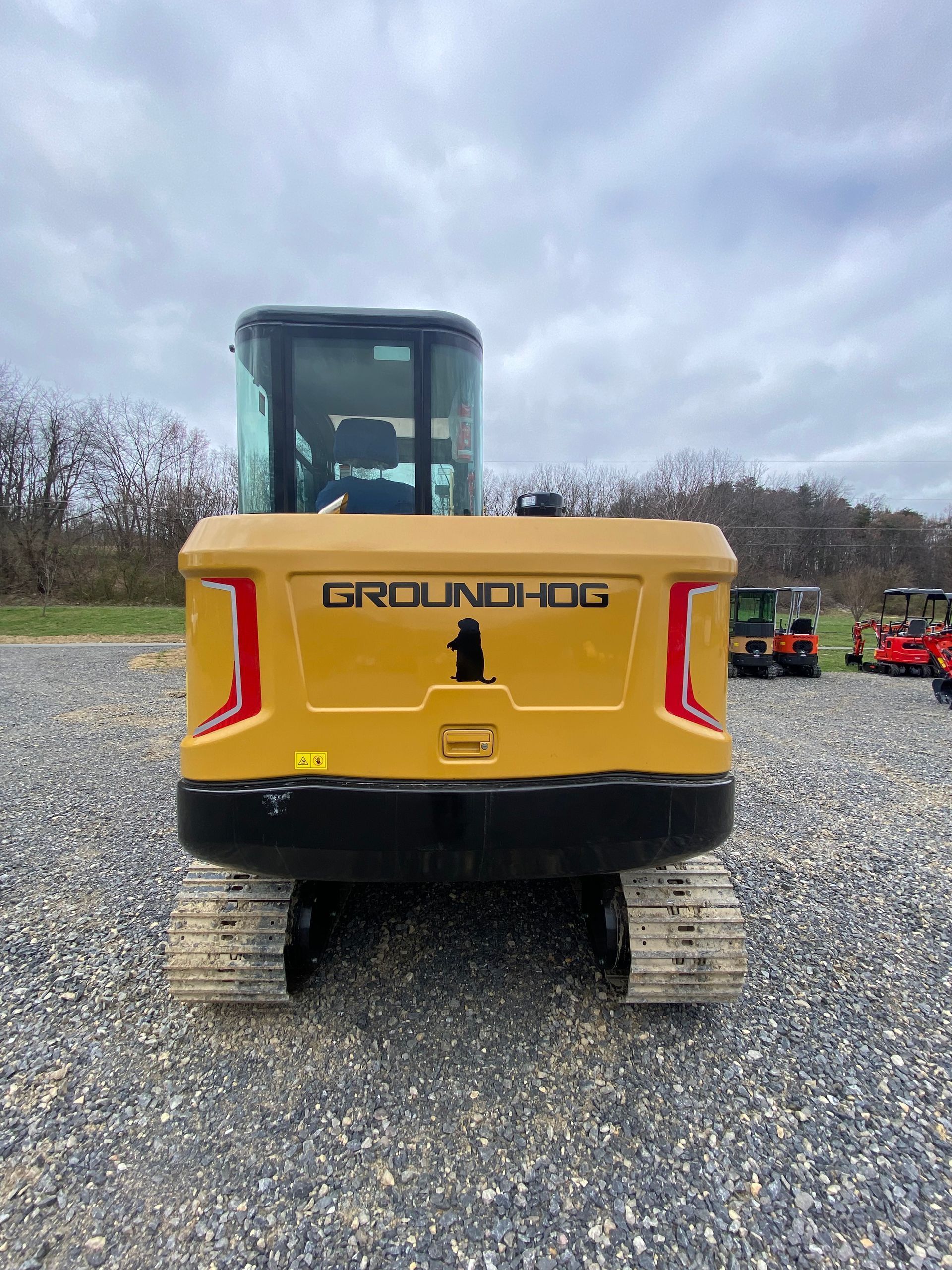 A rear view of a yellow Groundhog brand compact excavator parked on a gravel lot under a cloudy sky.